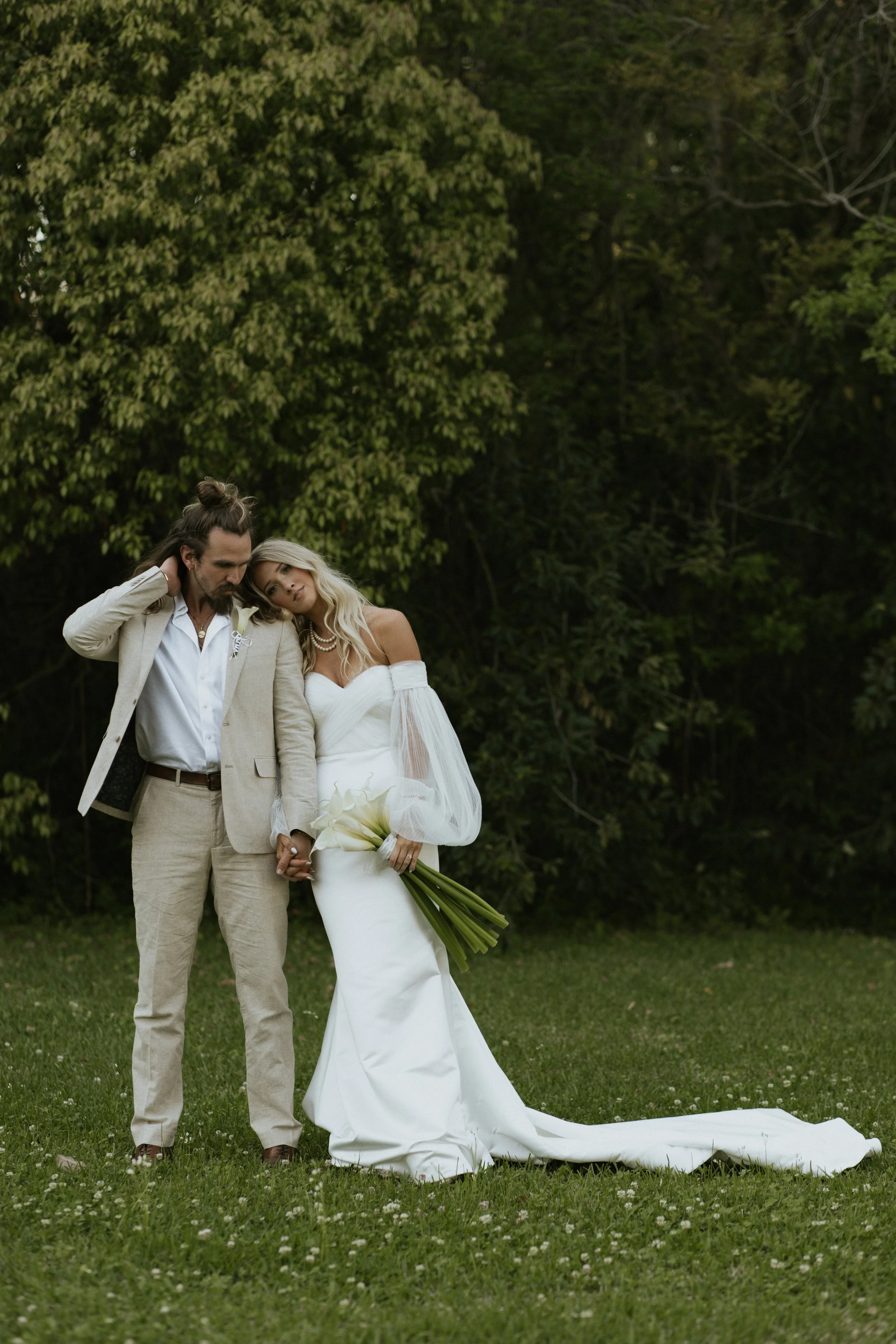 A bride and groom standing close together outdoors on grass with green trees in the background. The bride is holding a bouquet of calla lilies and the groom is touching his head with his hand, both with somber expressions.
