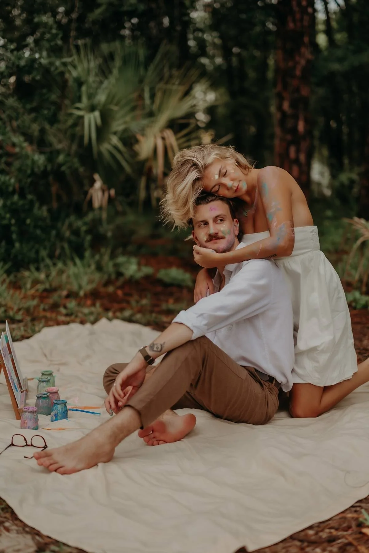 A couple sitting on a blanket in a forest, with the woman kneeling behind the man, embracing him, both with colorful paint on their bodies. The man has tattoos and glasses on the blanket, surrounded by small paint jars and brushes.