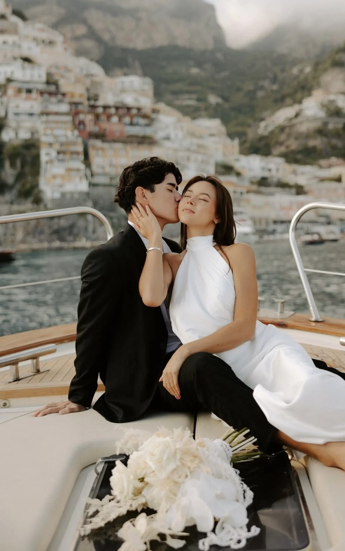 A couple sitting on a boat, with the man kissing the woman on the cheek as he holds her face. The woman is smiling with her eyes closed, dressed in a white dress. The man wears a black suit. The background features a harbor with colorful hillside houses and a mountain backdrop.