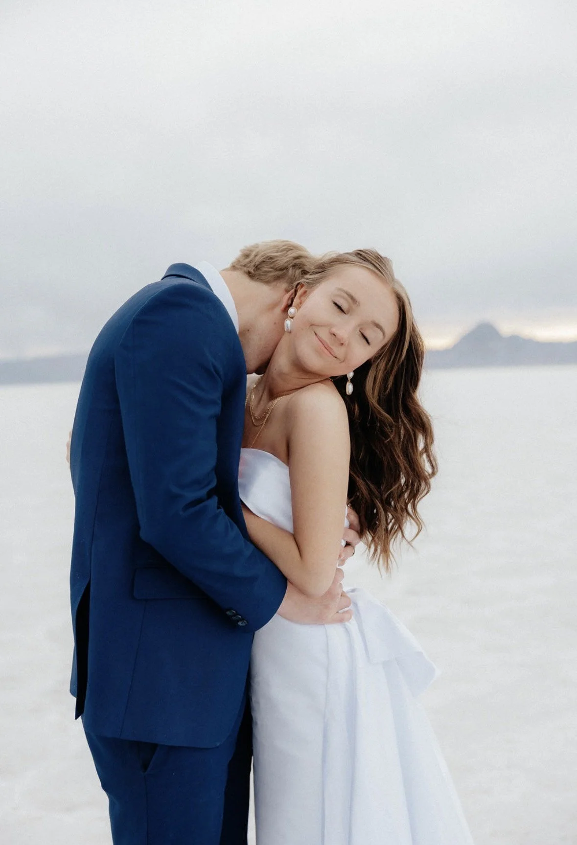 A woman in a white dress and a man in a blue suit embracing with their eyes closed on a beach with a mountain in the background.