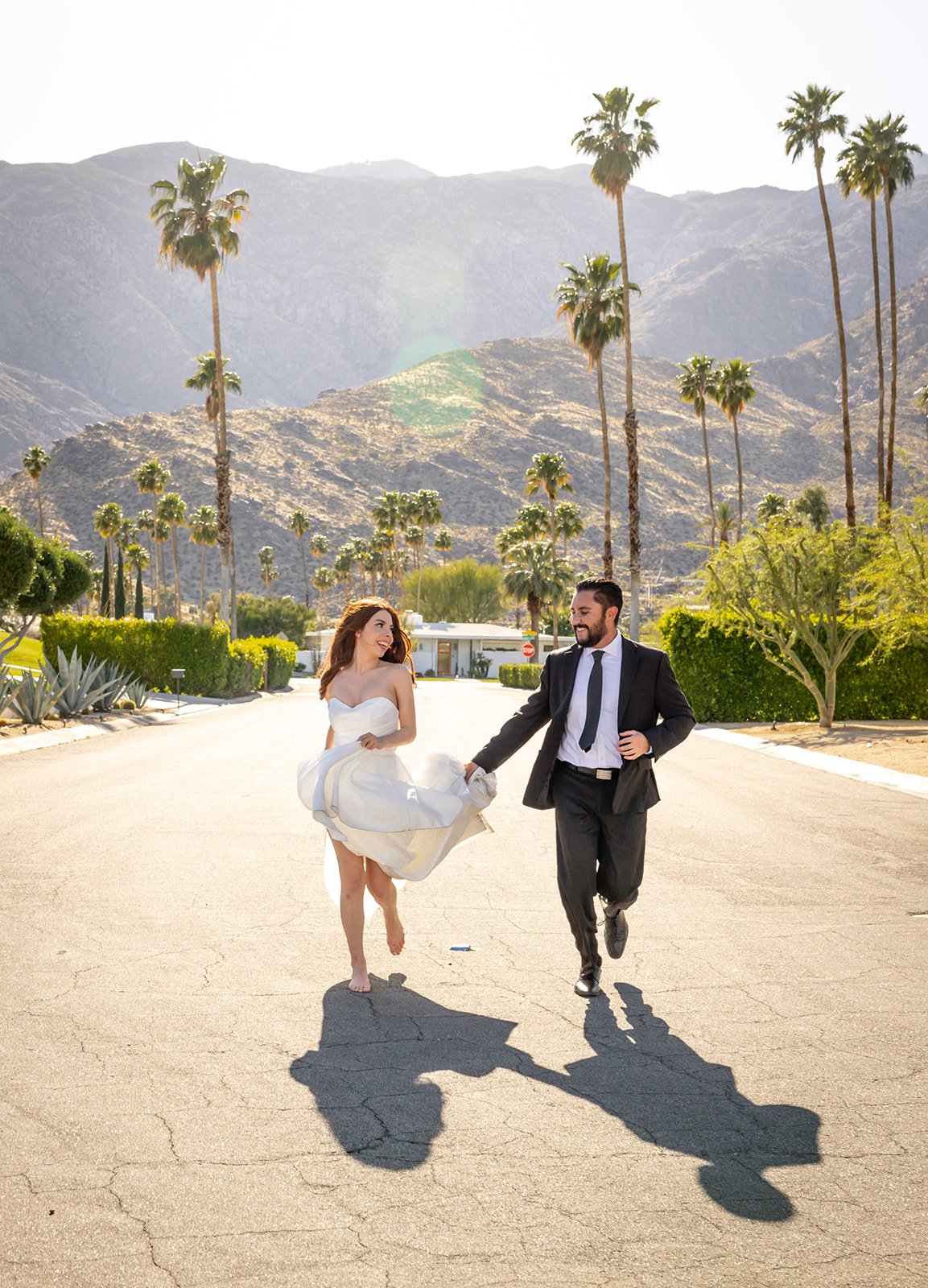 A newlywed couple holding hands and running barefoot on a street in a sunny, mountainous area with palm trees, wearing wedding attire. The bride is in a white dress, and the groom is in a black suit.