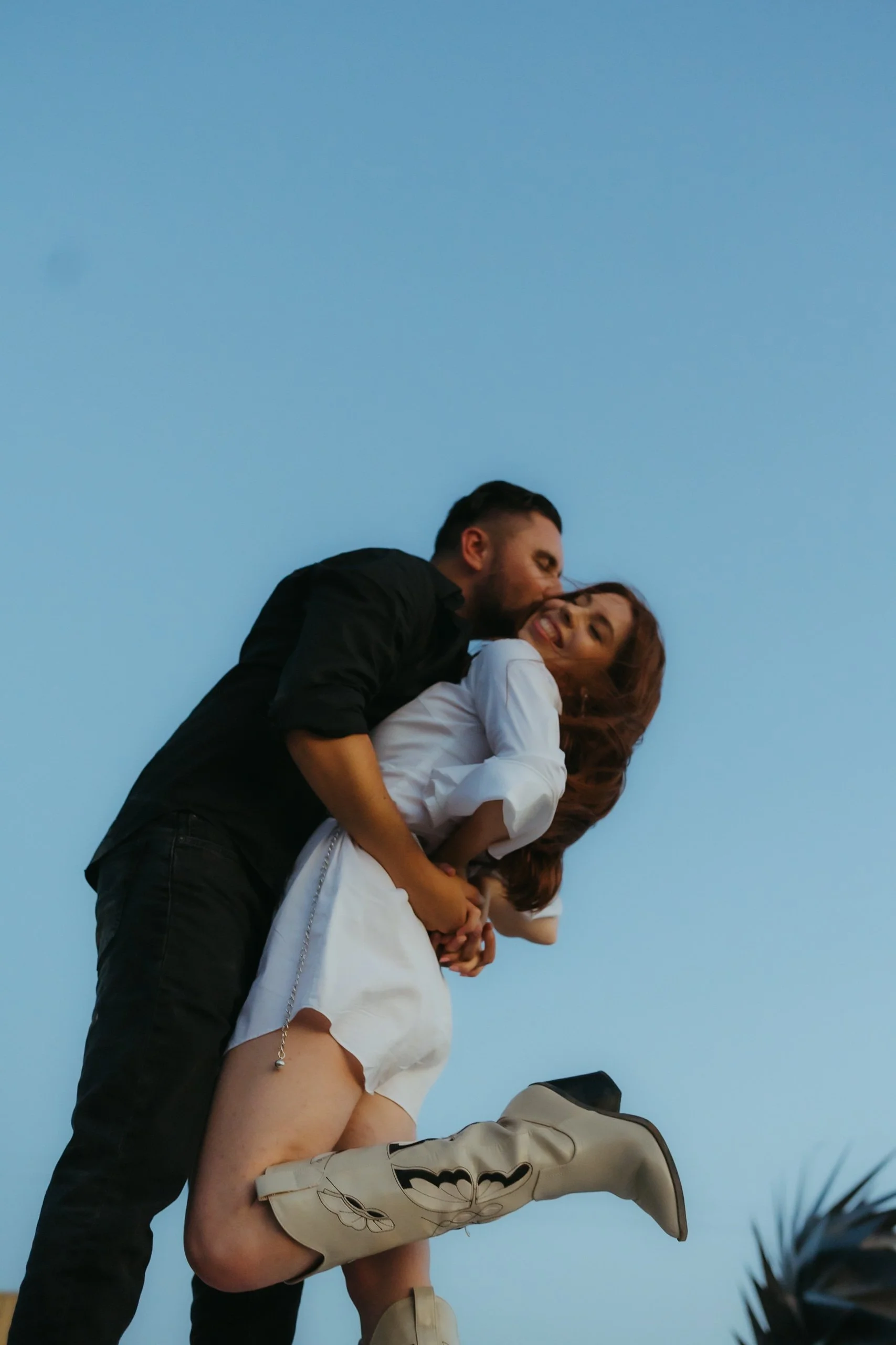 A couple sharing a romantic moment outdoors against a clear blue sky, with the man kissing the woman on the cheek. The woman is smiling and holding hands with the man, wearing cowboy boots.