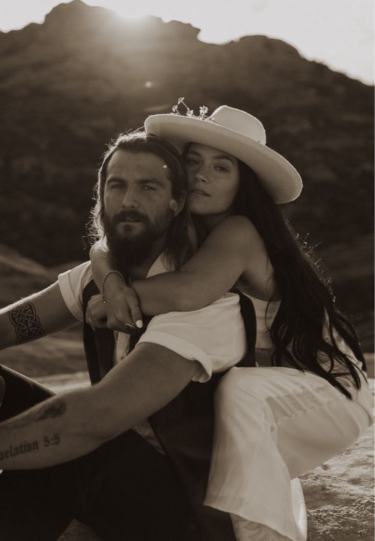 A black and white photo of a couple outdoors, with a mountain background, the woman hugging the man and wearing a wide-brimmed hat.