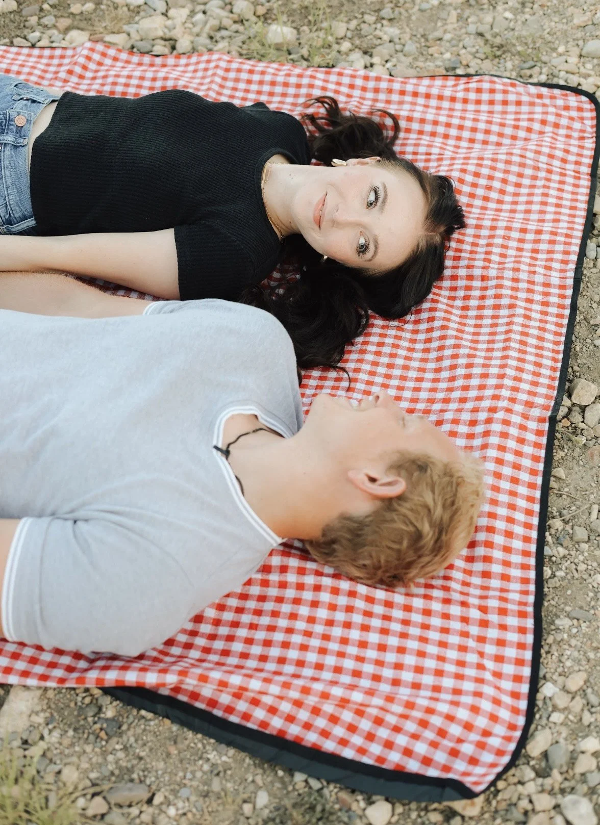 A woman with dark hair and a black top lying on a red and white checkered blanket; a man with blond hair and a gray shirt lying beside her. They are outdoors on rocky ground.