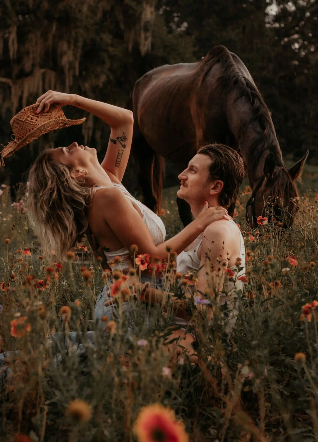 A woman and a man sitting in a field of flowers, with a horse grazing nearby in a natural, outdoor setting at sunset.