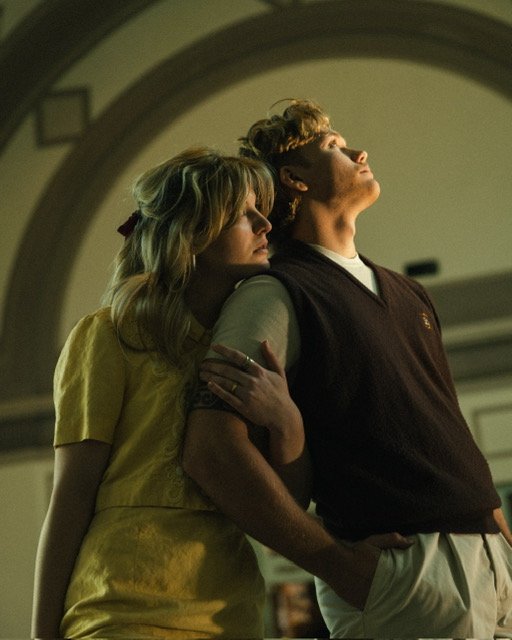A young woman leaning on a young man, both with eyes closed, inside a church or hall with arched ceiling.