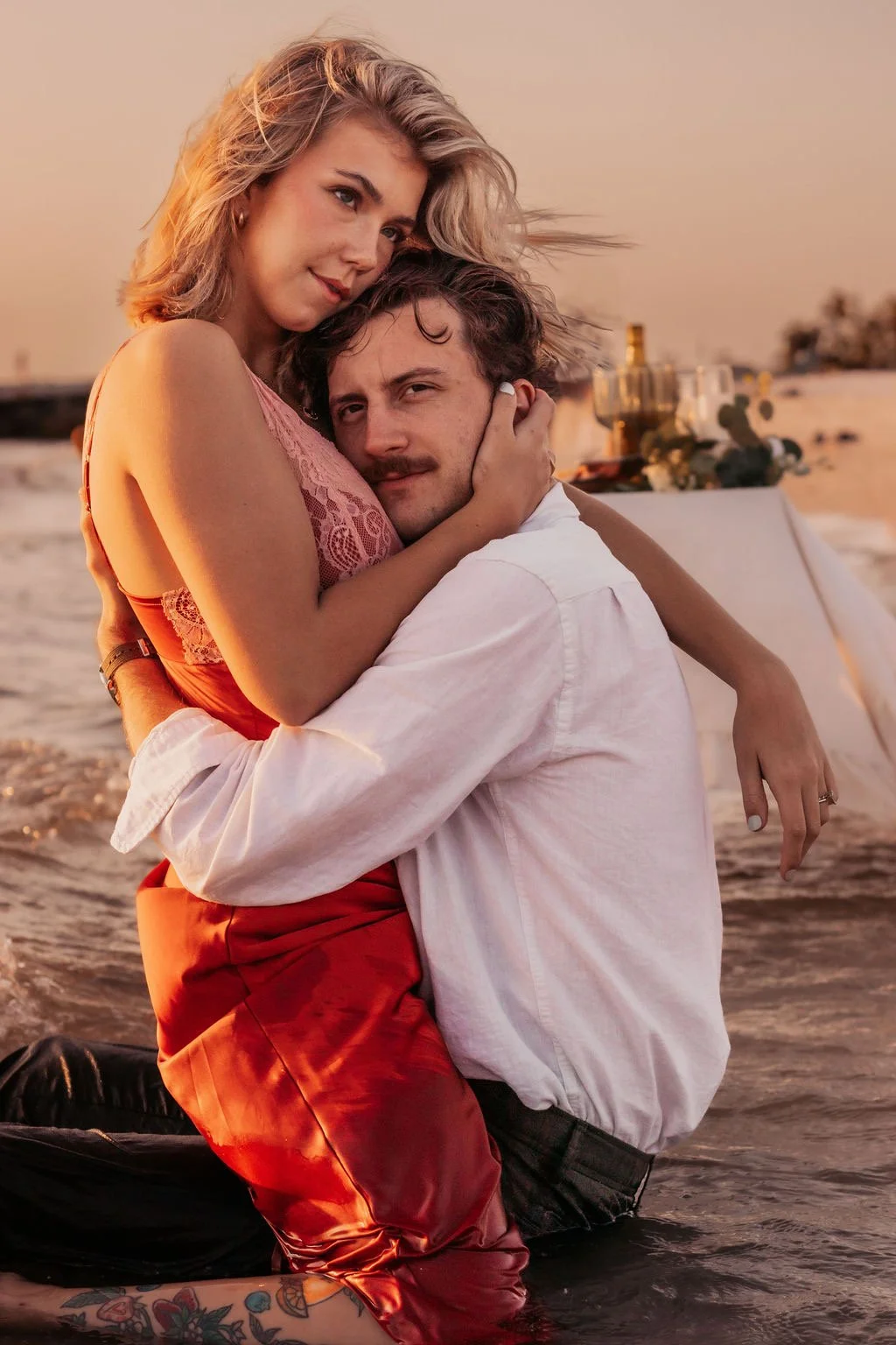 A couple embracing in shallow water on the beach during sunset, with a table set for a romantic dinner in the background.