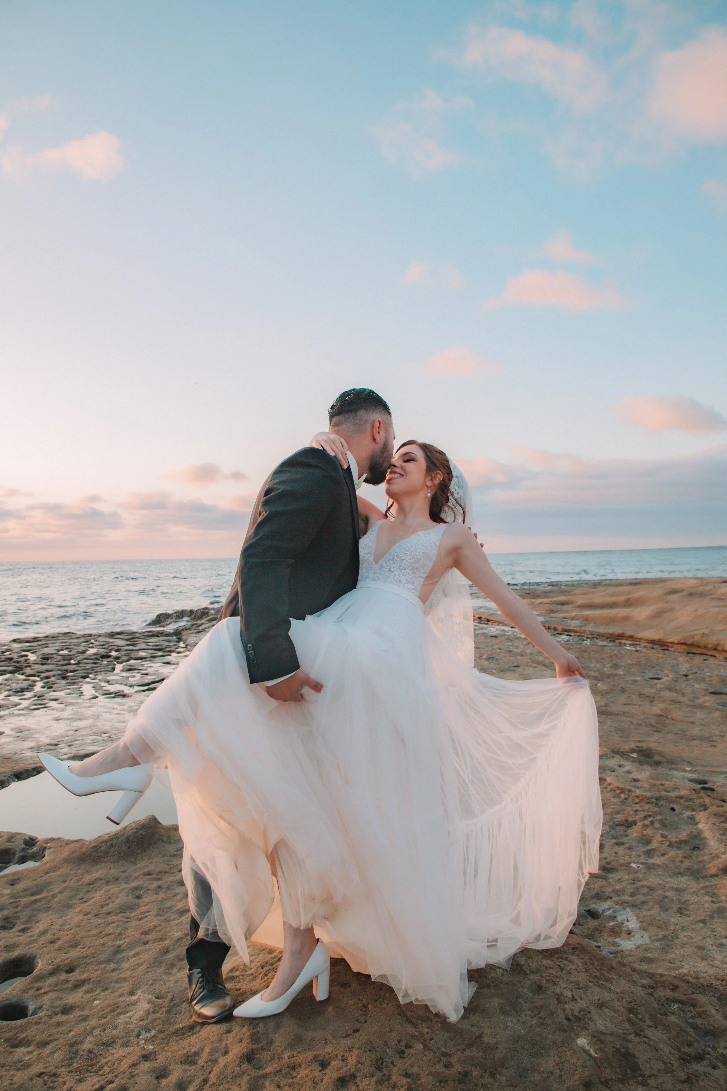 A newlywed couple dance on the beach during sunset, with the groom holding the bride and both smiling.