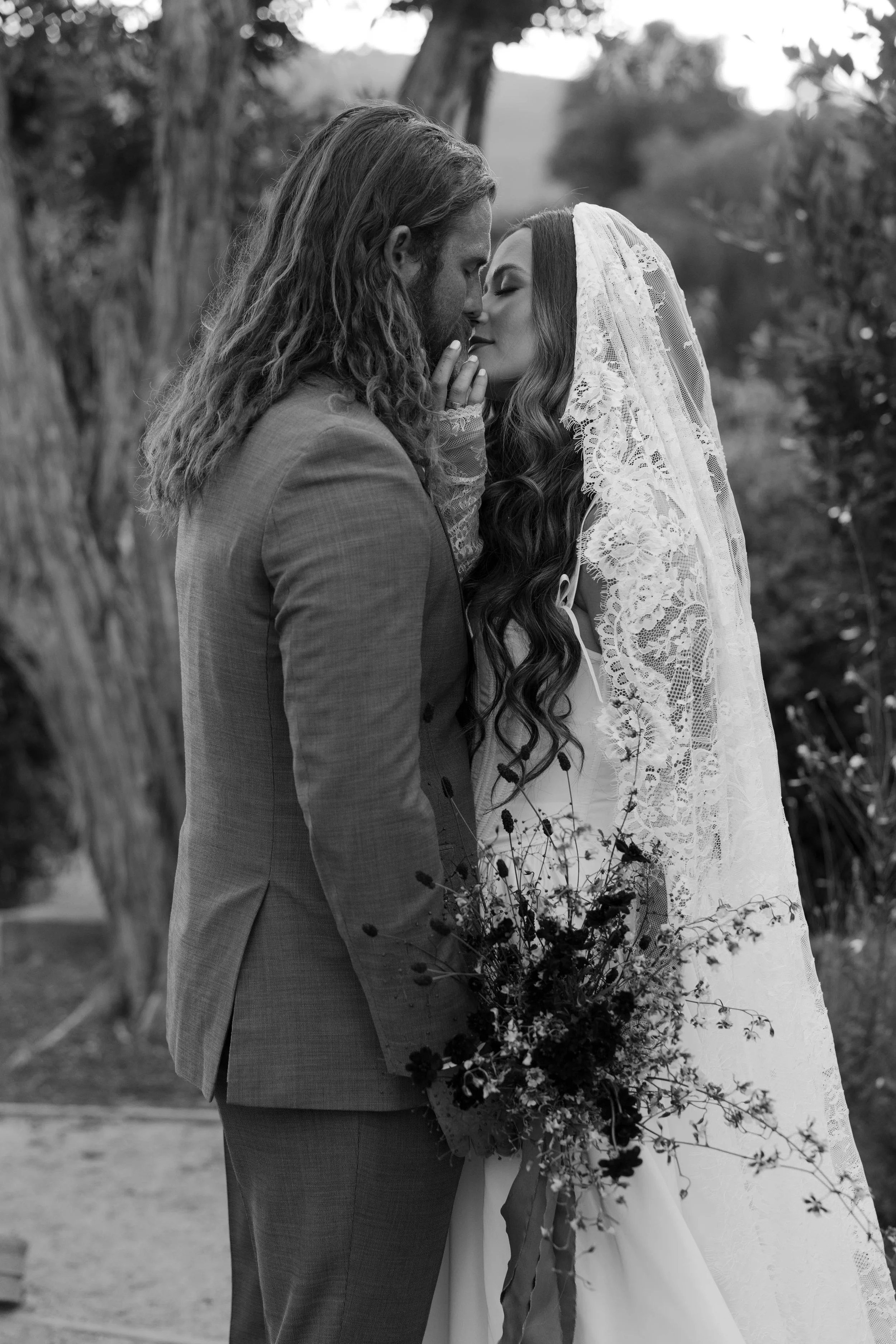 A black and white photo of a bride and groom in an outdoor setting, close together with foreheads touching, the bride holding a bouquet, and both in romantic poses.