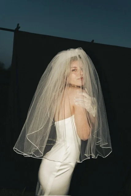 Woman in a white dress with blonde hair, wearing a veil and gloves, posing against a dark background.