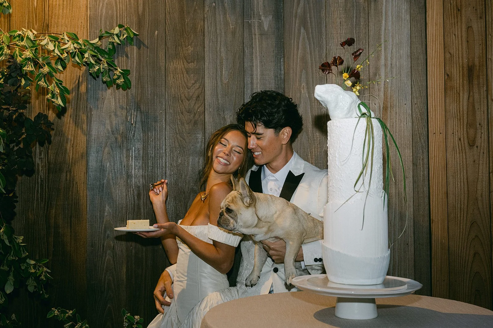 A couple celebrating with a small dog and cake in front of a wooden wall, with a large white cake decorated with flowers in the foreground.