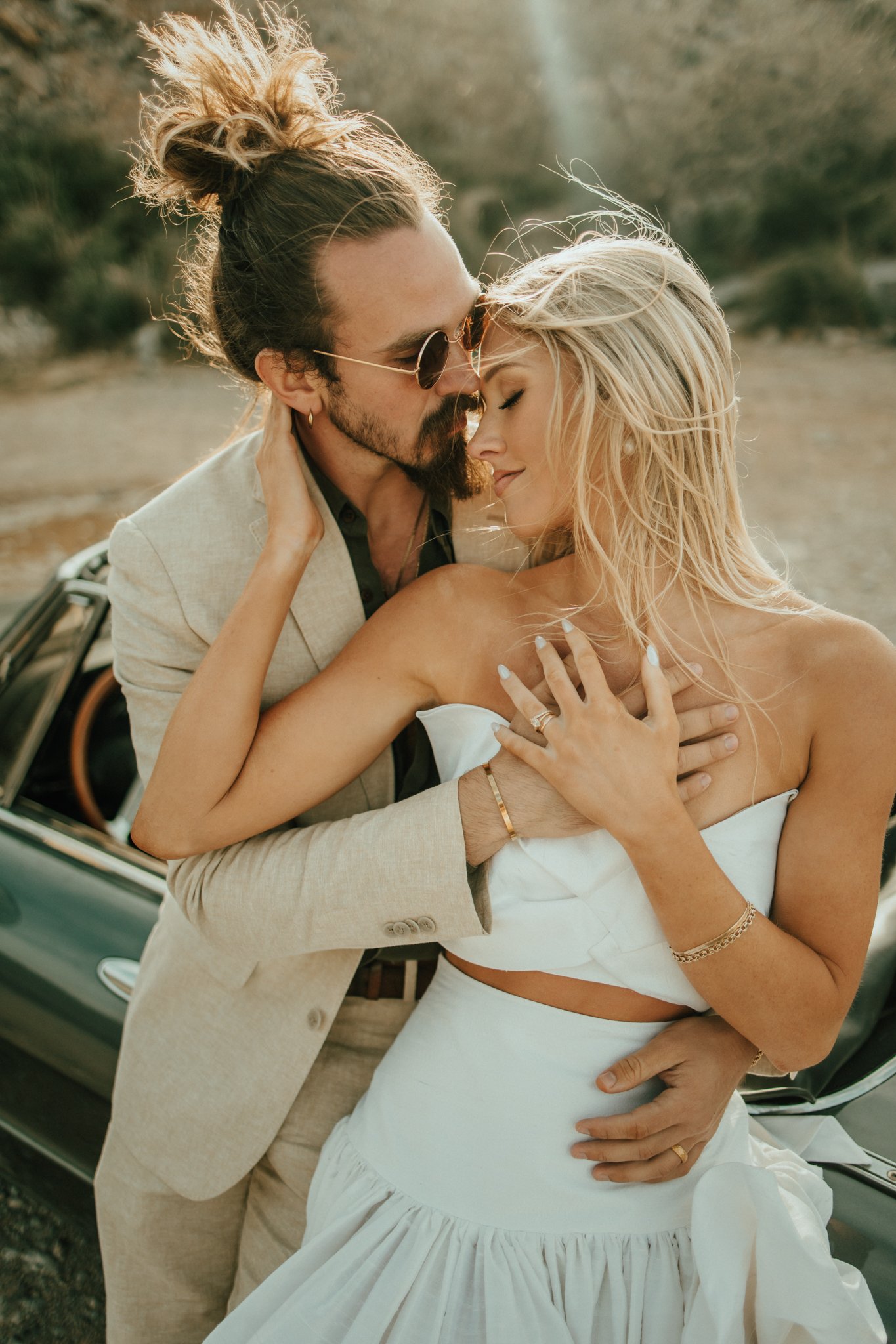 A couple embracing outdoors, with the man wearing sunglasses and a beige suit and the woman in a white dress, both with their eyes closed, softly touching and leaning into each other, in a warm, golden light setting.