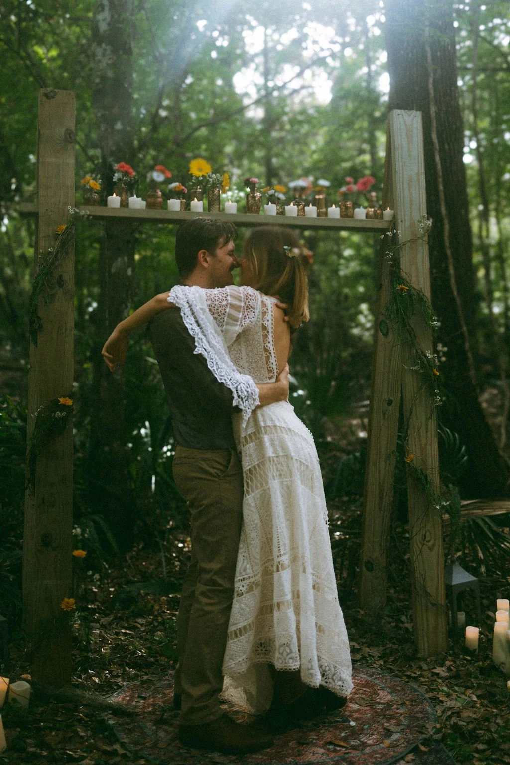A couple embracing in a forest wedding ceremony with a wooden arch decorated with candles and flowers.