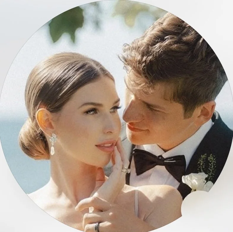 A bride and groom in wedding attire, close together outdoors, with the bride holding her finger to her lips and the groom smiling