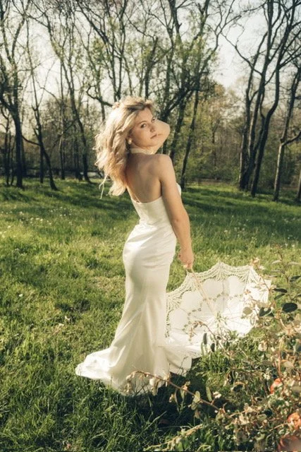 A woman in a white dress standing in a grassy field with trees in the background, holding an open lace parasol.