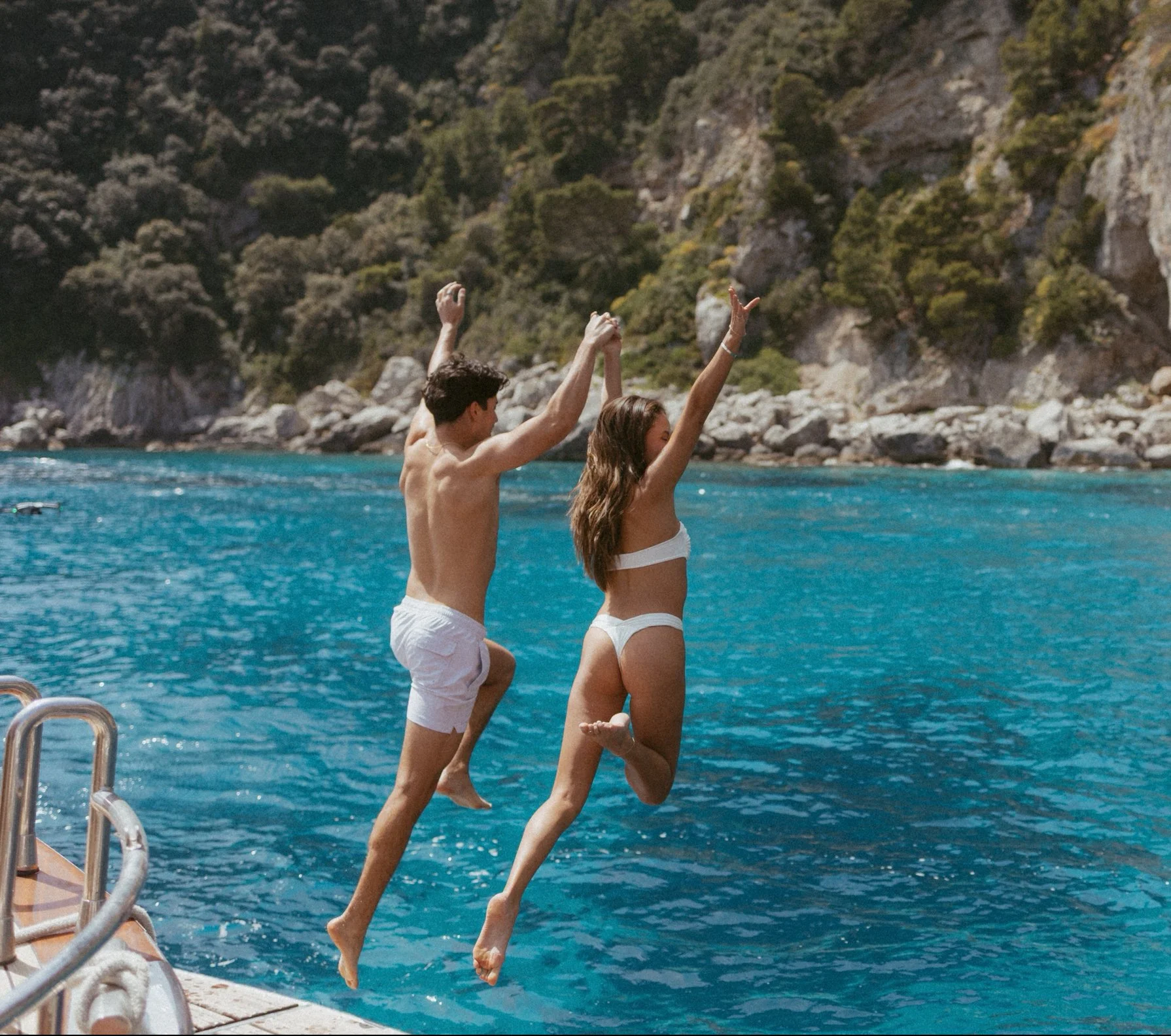 A couple in swimsuits jumping into water from a dock, with a rocky coastline and green hills in the background.