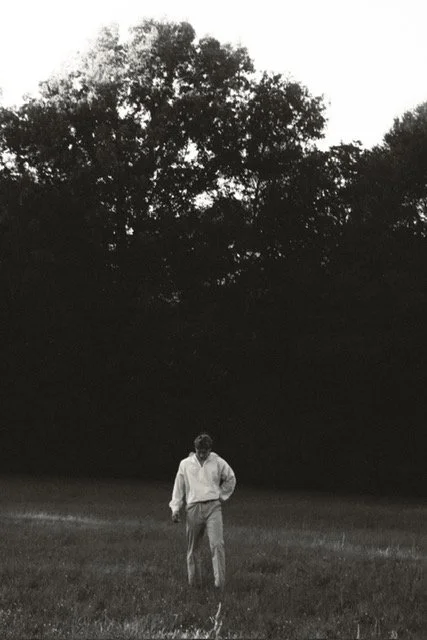 A person walking on grass in front of tall trees, with dark foliage and sky in the background, in a black and white photograph.