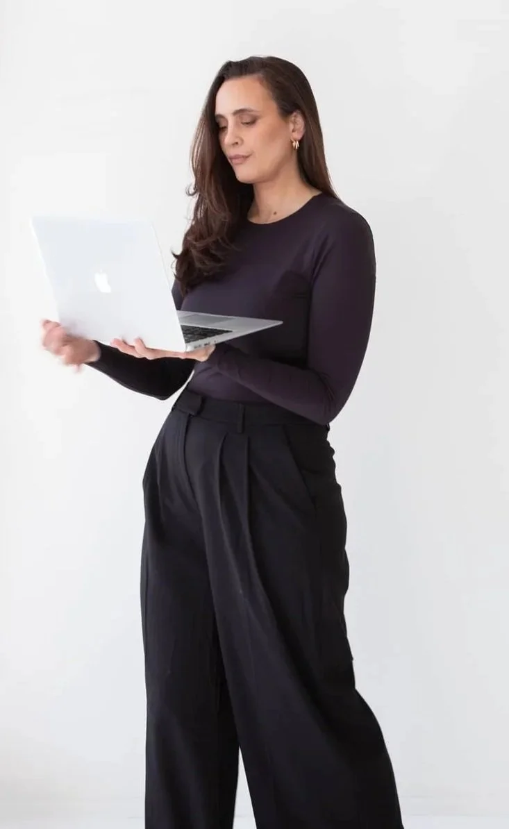 Angelique from Kitsa Creative in black long-sleeve top and high-waisted black pants holding and looking at a silver Apple MacBook against a plain white background.