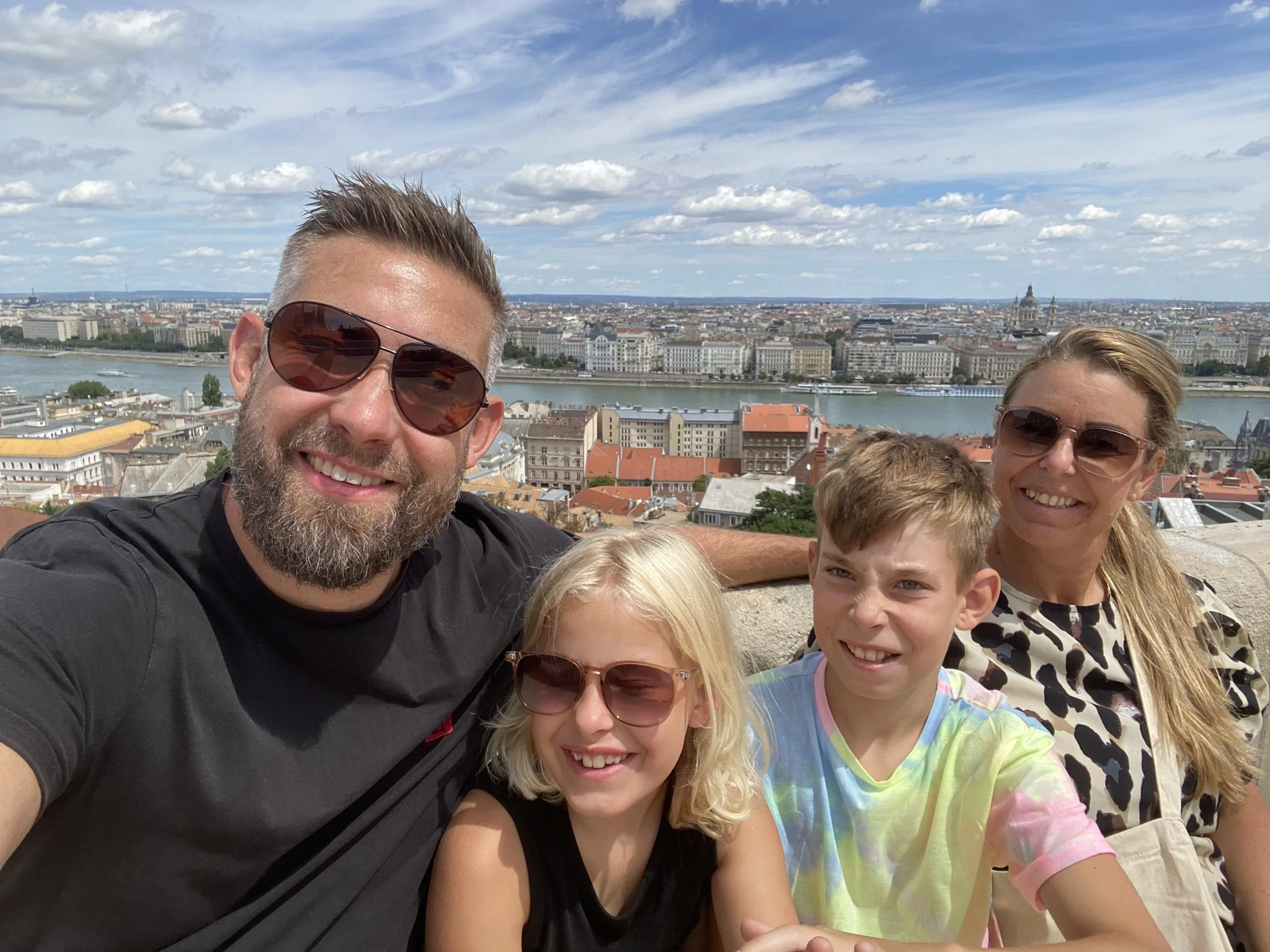 En familie på fire tager selfie med en bys skyline og en flod i baggrunden, under en blå himmel med spredte skyer.