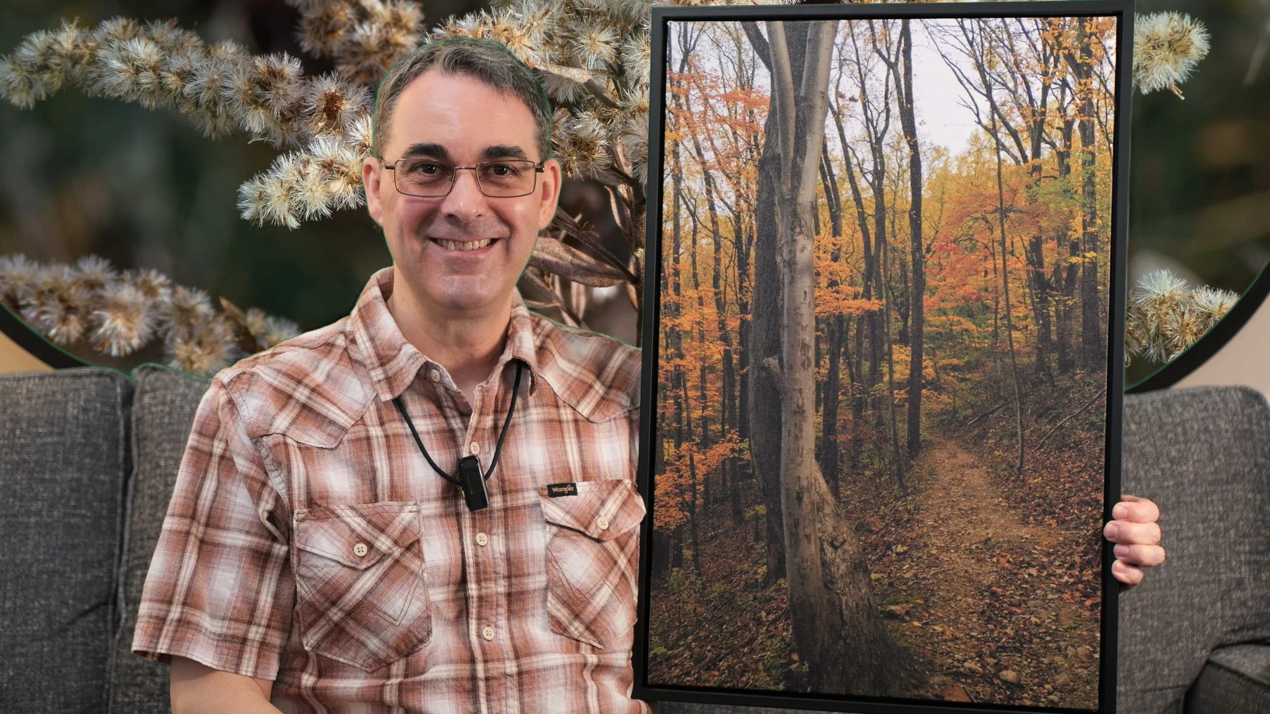 The photographer is sitting on a couch showing a canvas photo print of a forest trail under orange fall foliage and an overcast sky.