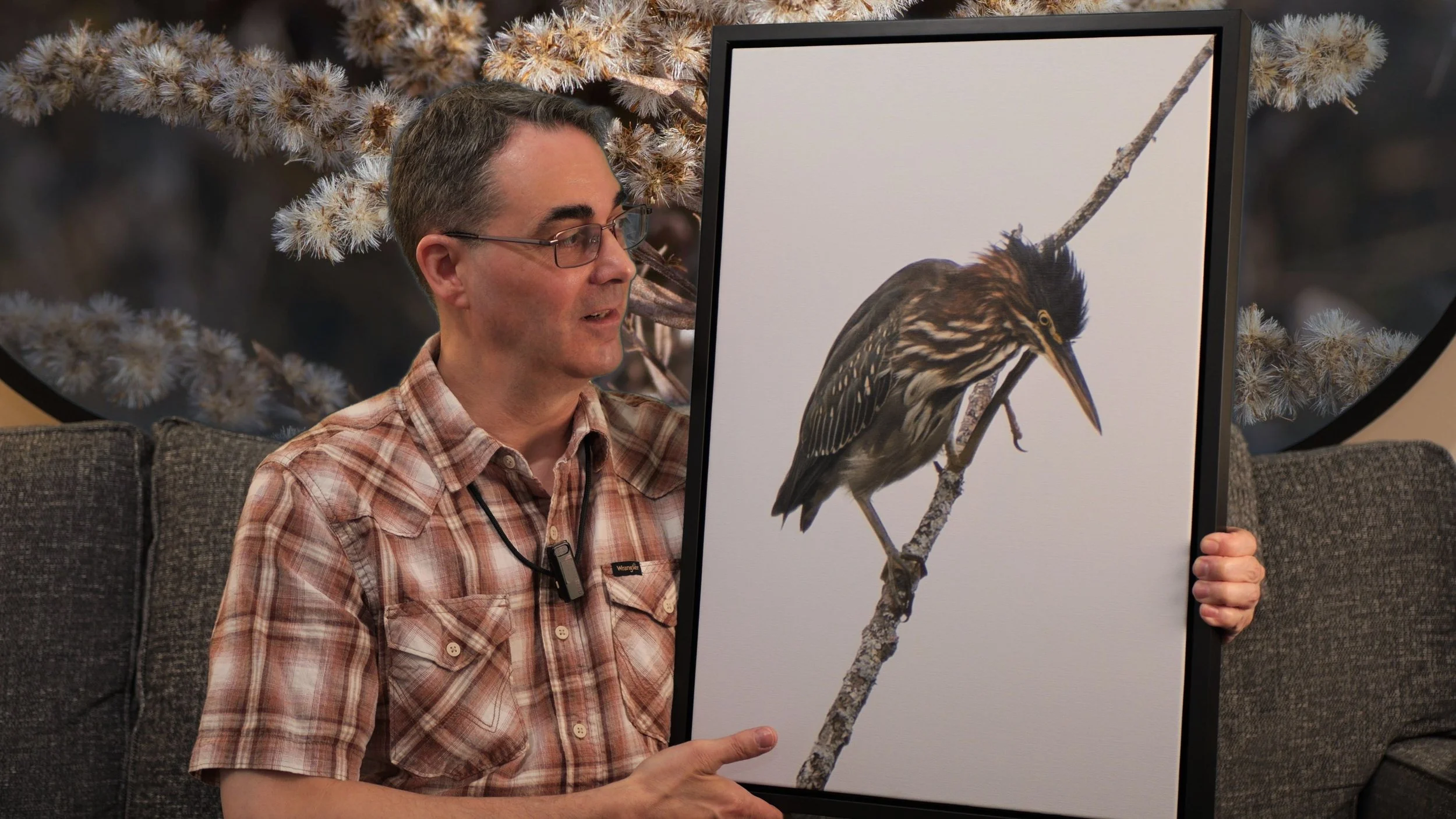 The photographer is sitting on a couch showing a canvas photo print of a green heron scratching its head while perched on a branch with a white background.