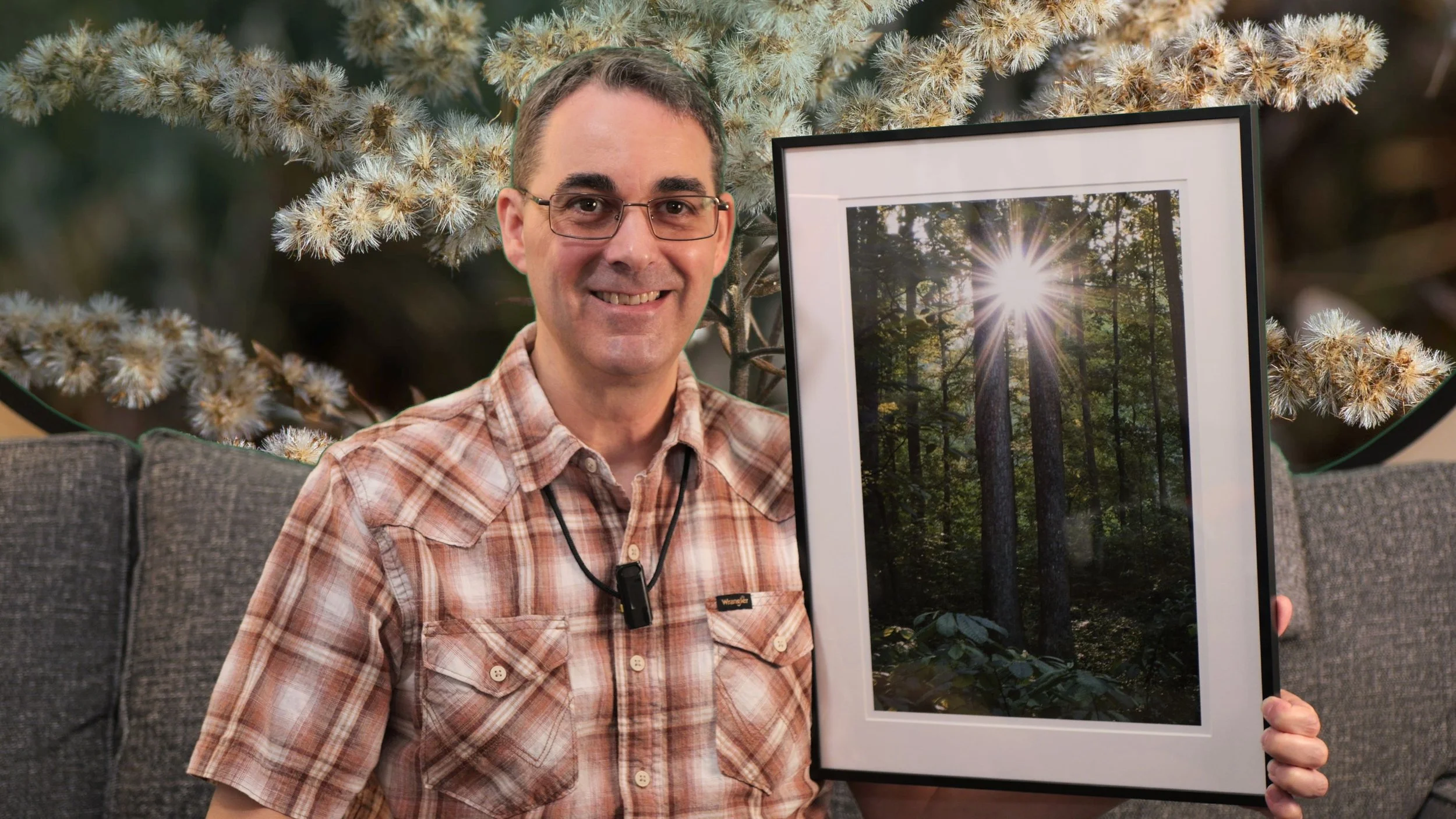 The photographer is sitting on a couch showing a framed photo of a sunburst shining through large twin trees in a forest.