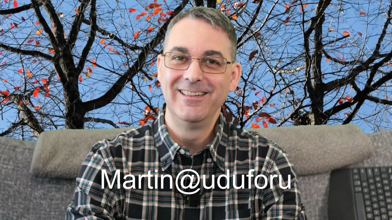 A photo of Martin from the shoulders up, smiling and sitting on a couch. He is wearing a long-sleeve black and white flannel shirt, the couch is grey, and the wall behind is an image of black bare tree branches with sparse red leaves with a blue sky.