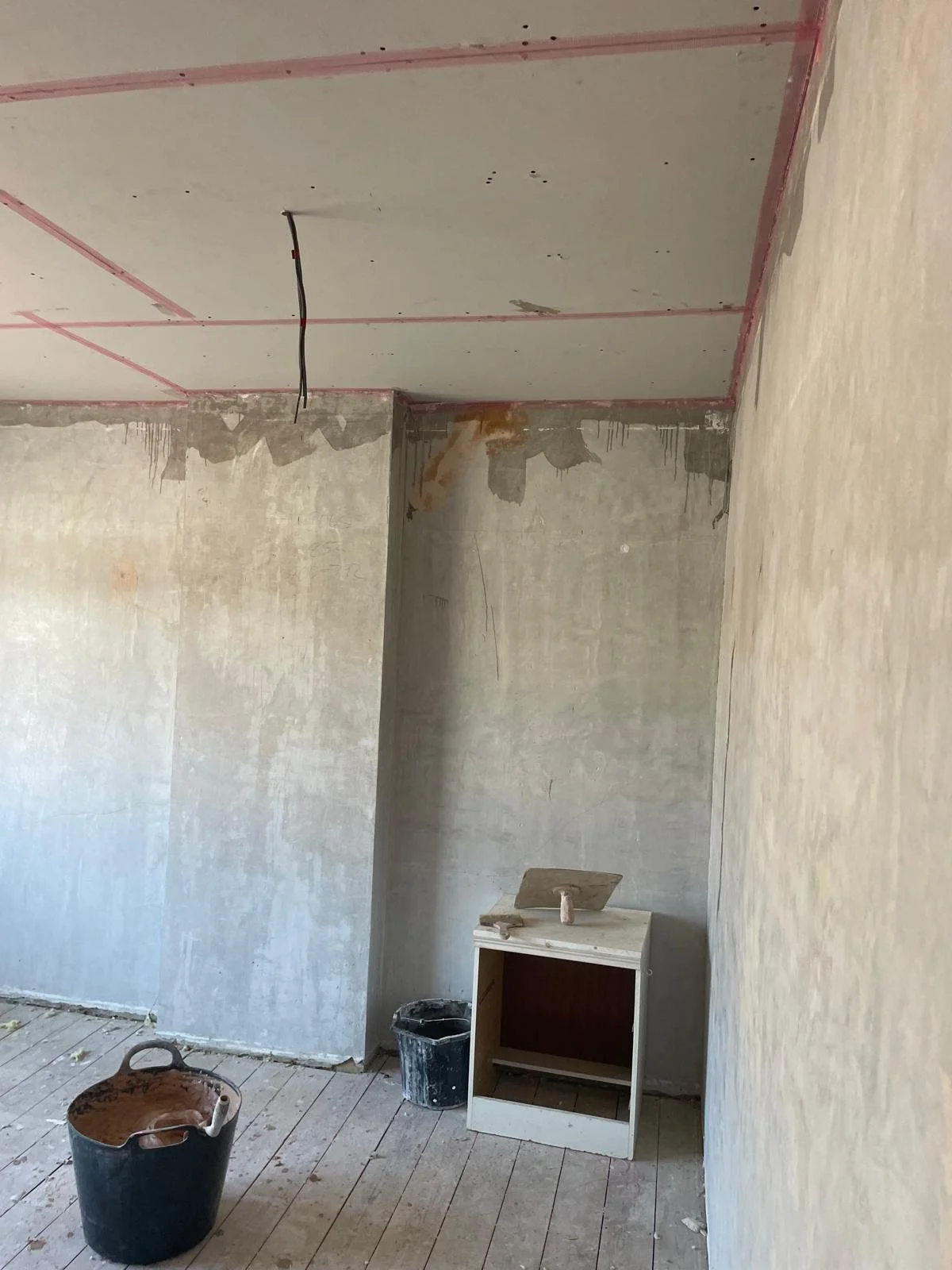 Room under construction with unfinished drywall and a small white cabinet, with a bucket of mud and construction tools on the floor