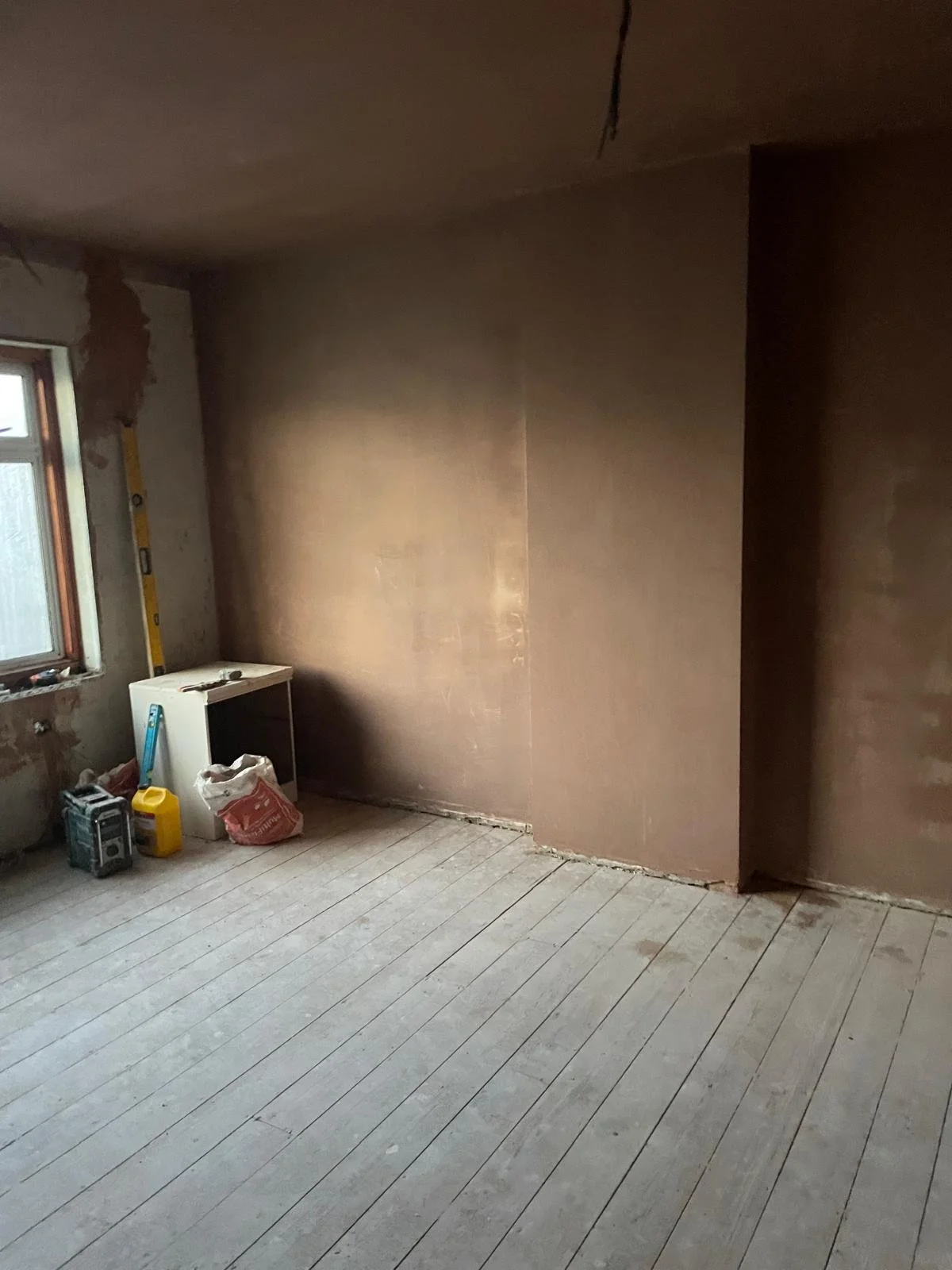 Room under renovation with unfinished walls and flooring, construction tools and supplies near the window.