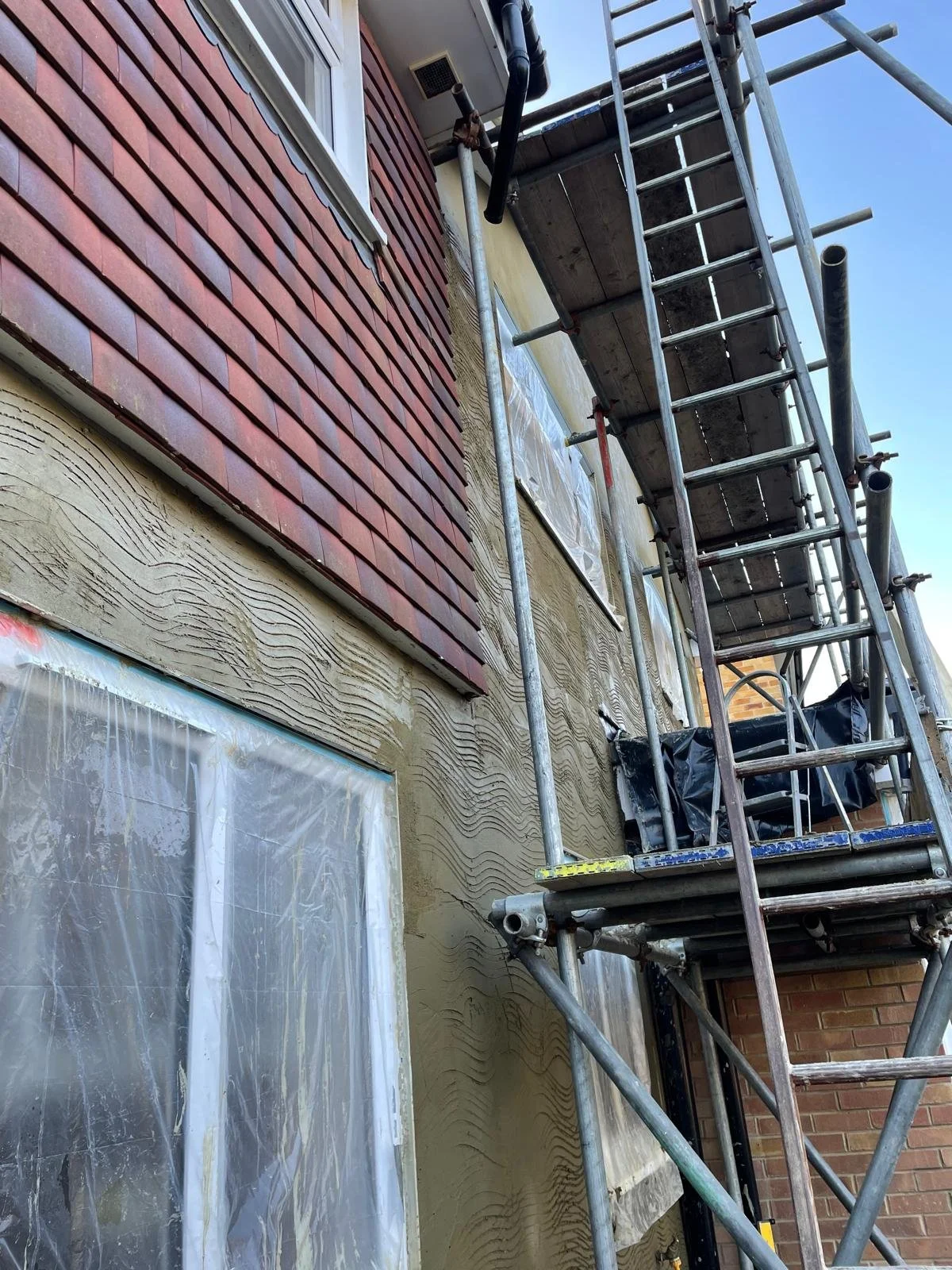 A house exterior under construction with scaffolding, showing partially finished wall with textured beige finish, a window covered with plastic sheeting, and a section of red shingle siding.