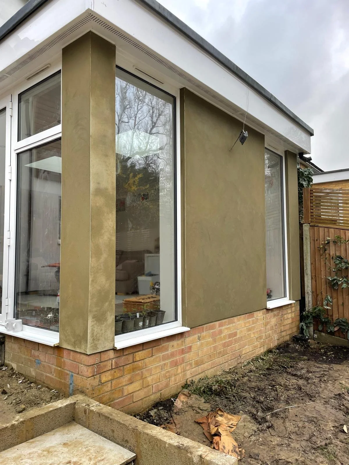 The exterior corner of a house with large windows, a brick foundation, and new greenish plastered wall panels, with some construction mud and materials in the front yard.