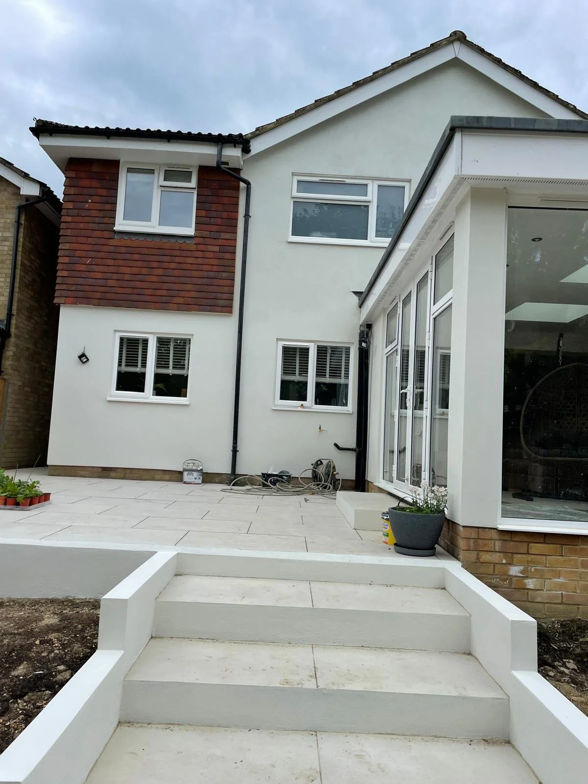 Back of a modern house with white exterior walls, large windows, a glass door, and newly laid patio with steps, under an overcast sky.