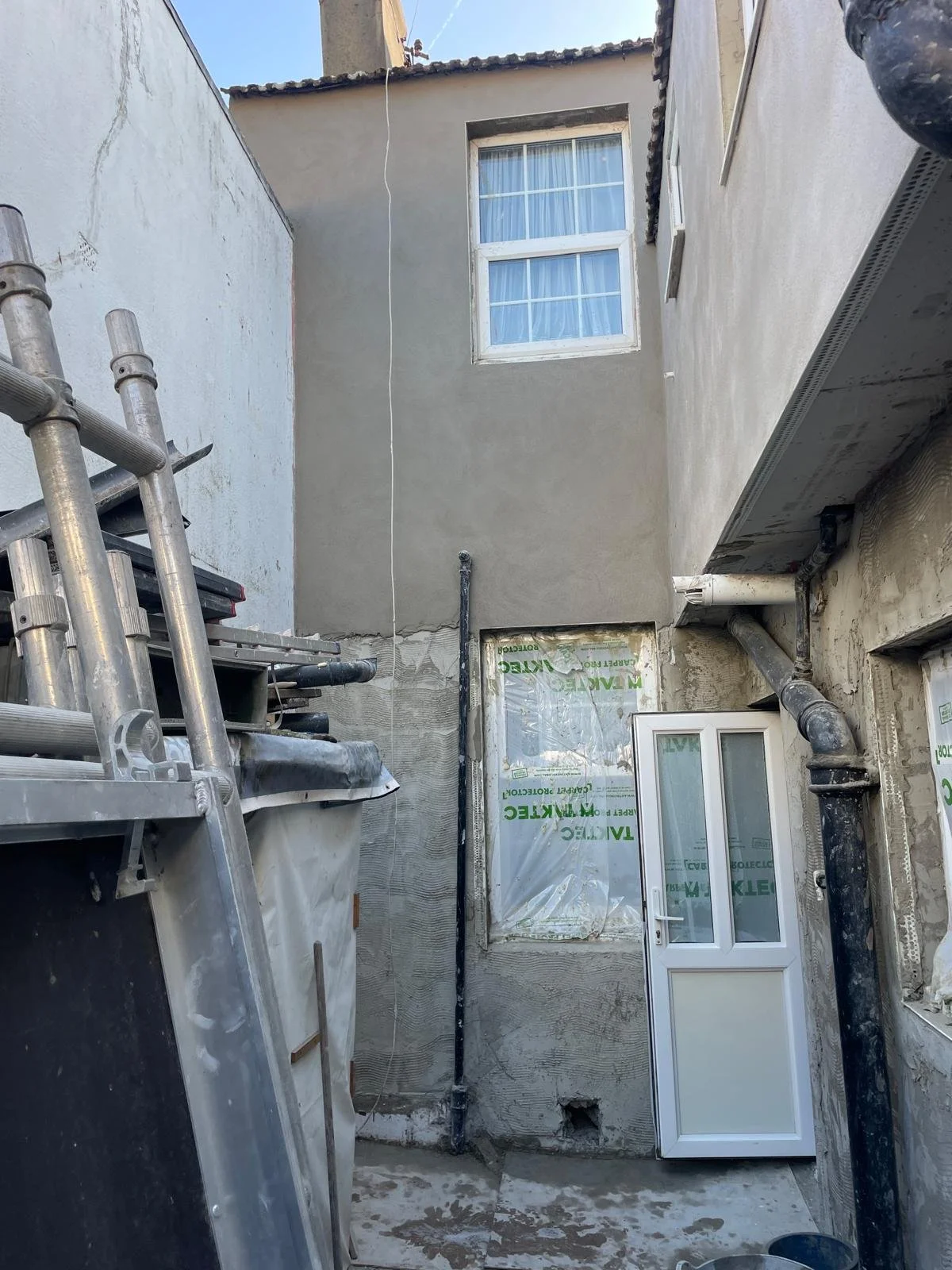 Building construction site with a partially finished wall, a small window, construction materials, pipes, and a white door.