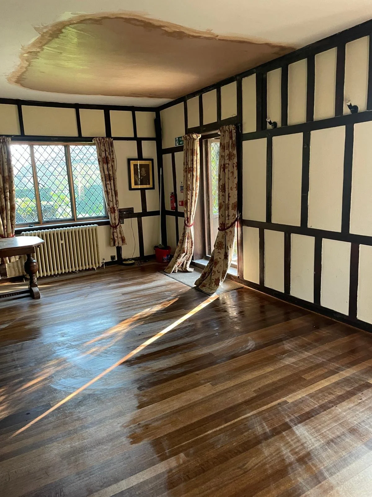 Empty room with wooden floor, window with diamond grid and floral curtains, a door with floral curtains, a radiator, and a ceiling with water damage.