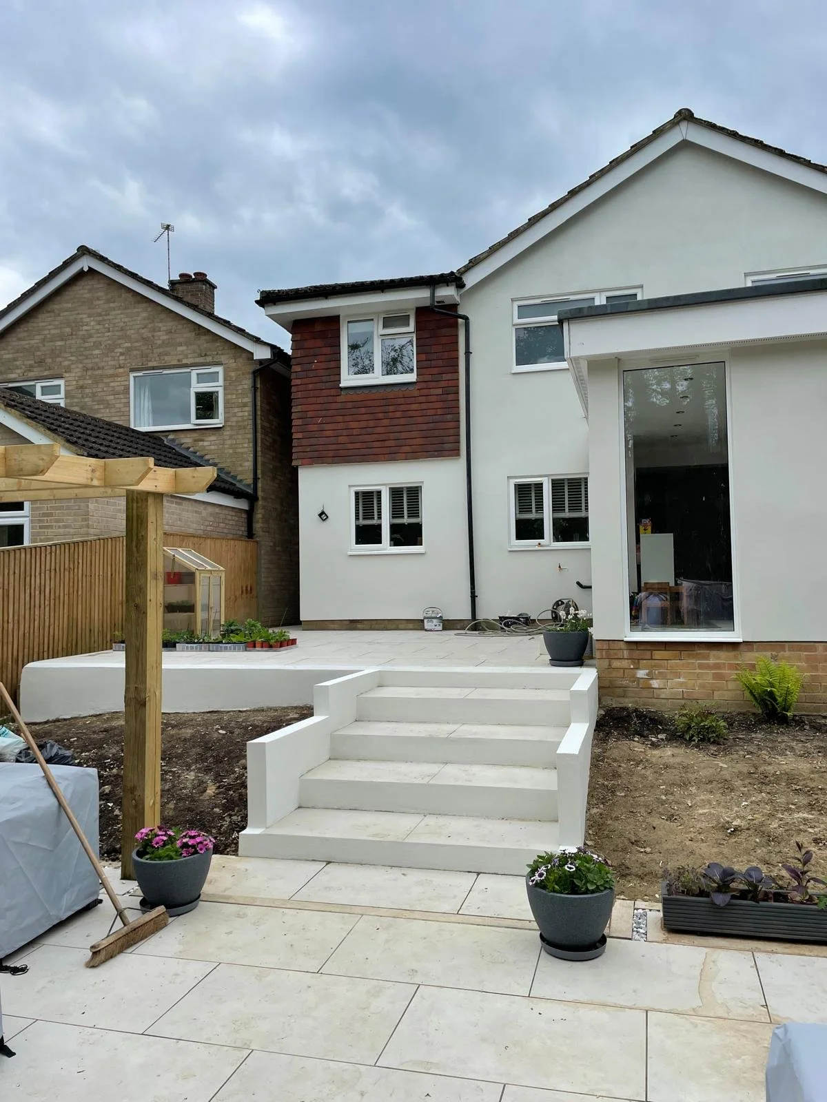 Backyard of a house under construction, with a white staircase leading to a patio, potted plants, and an unfinished garden area.