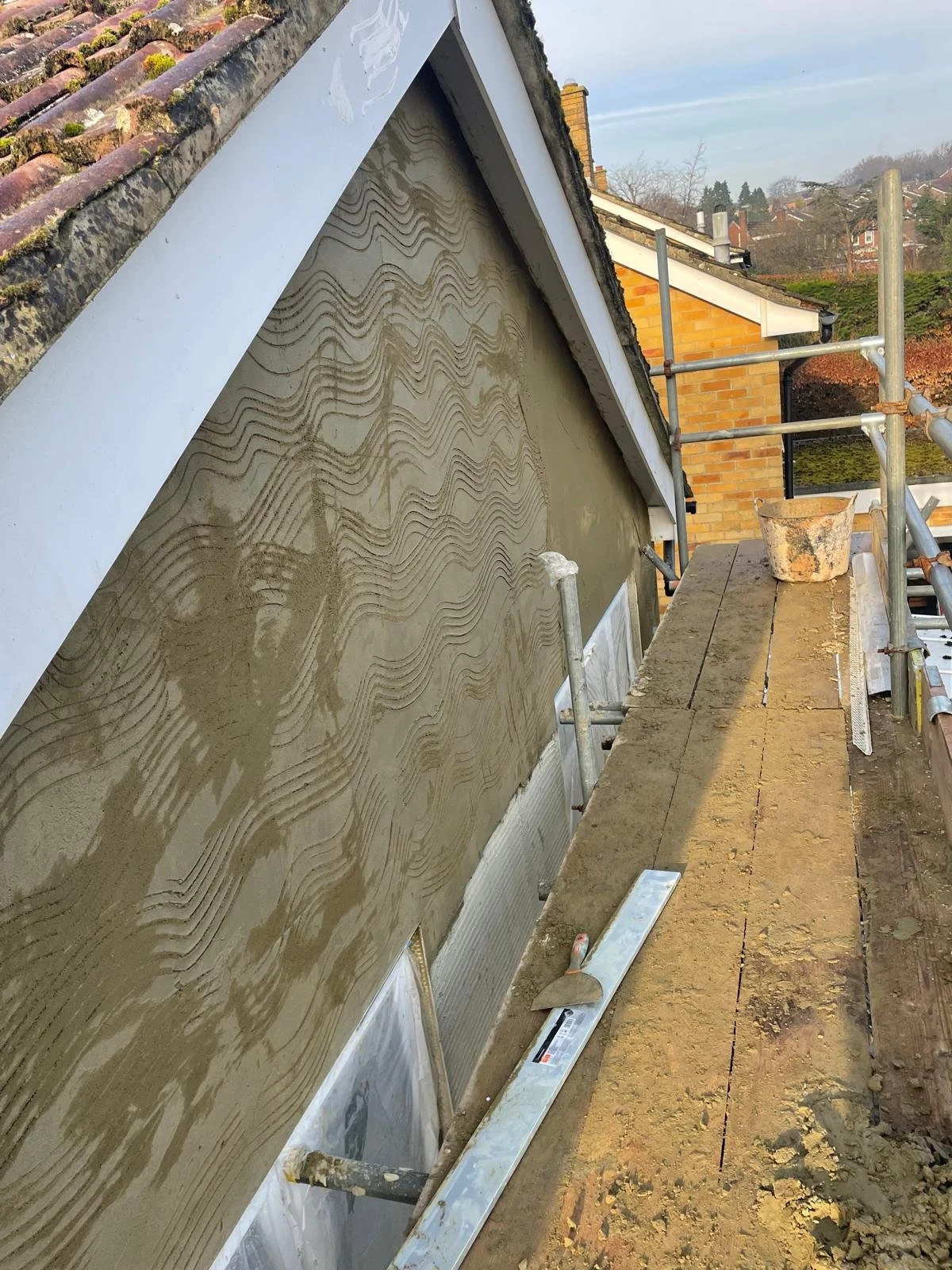 Construction site on a house roof with scaffolding, a newly coated wall with decorative sand texture, and a trowel resting on a wooden plank.