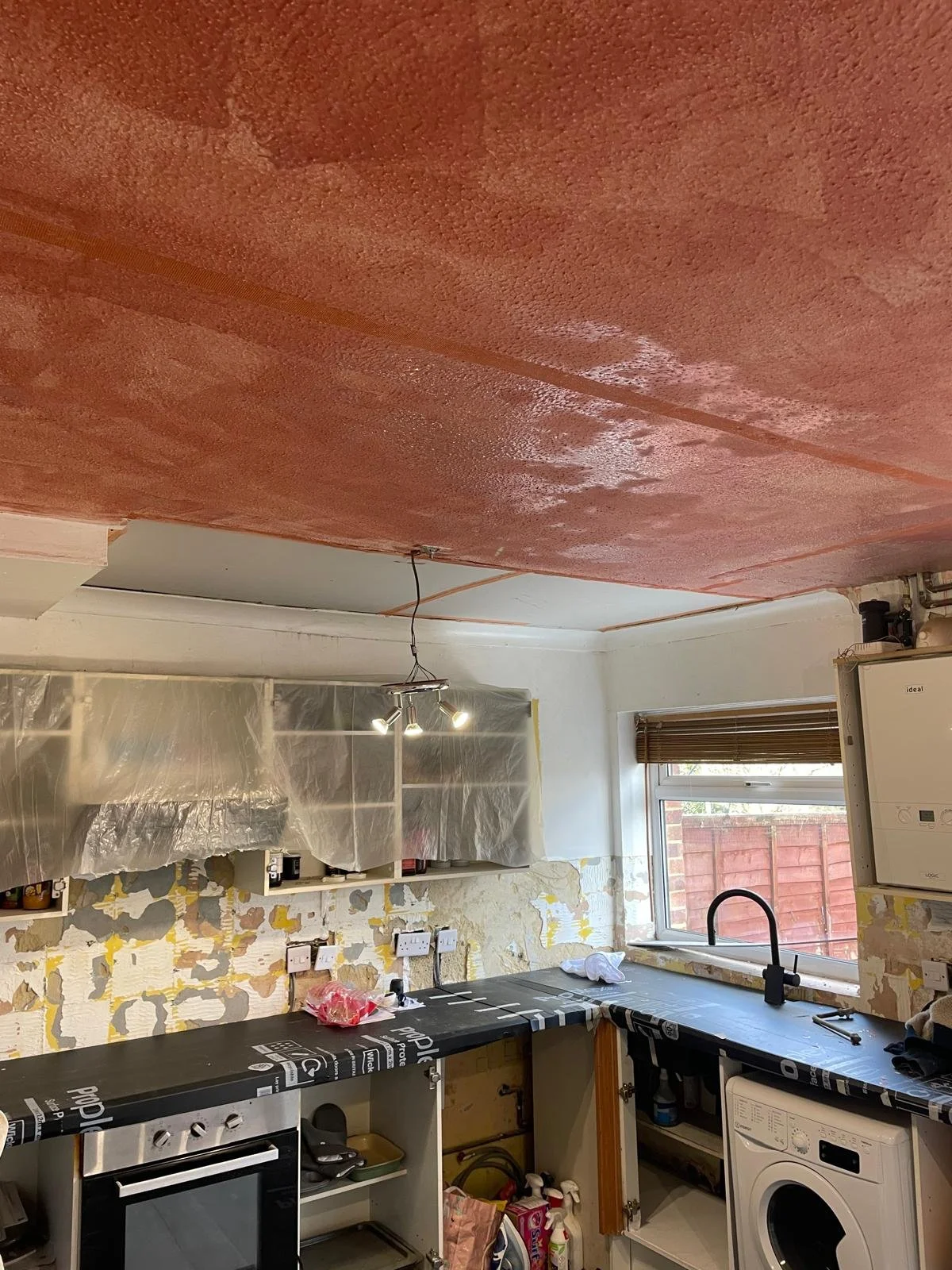 A kitchen ceiling painted in a reddish or pinkish color, with some painter's tape and plastic coverings on cabinets being prepared for painting. The room has a window with wooden blinds, and various kitchen appliances and cabinets in disarray, indica