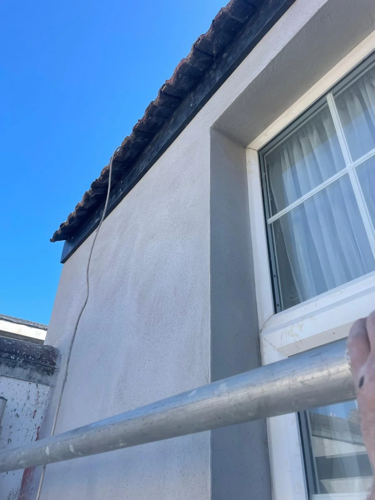 Close-up of a house exterior showing the corner of the wall, window, and roof with scaffolding in the foreground.