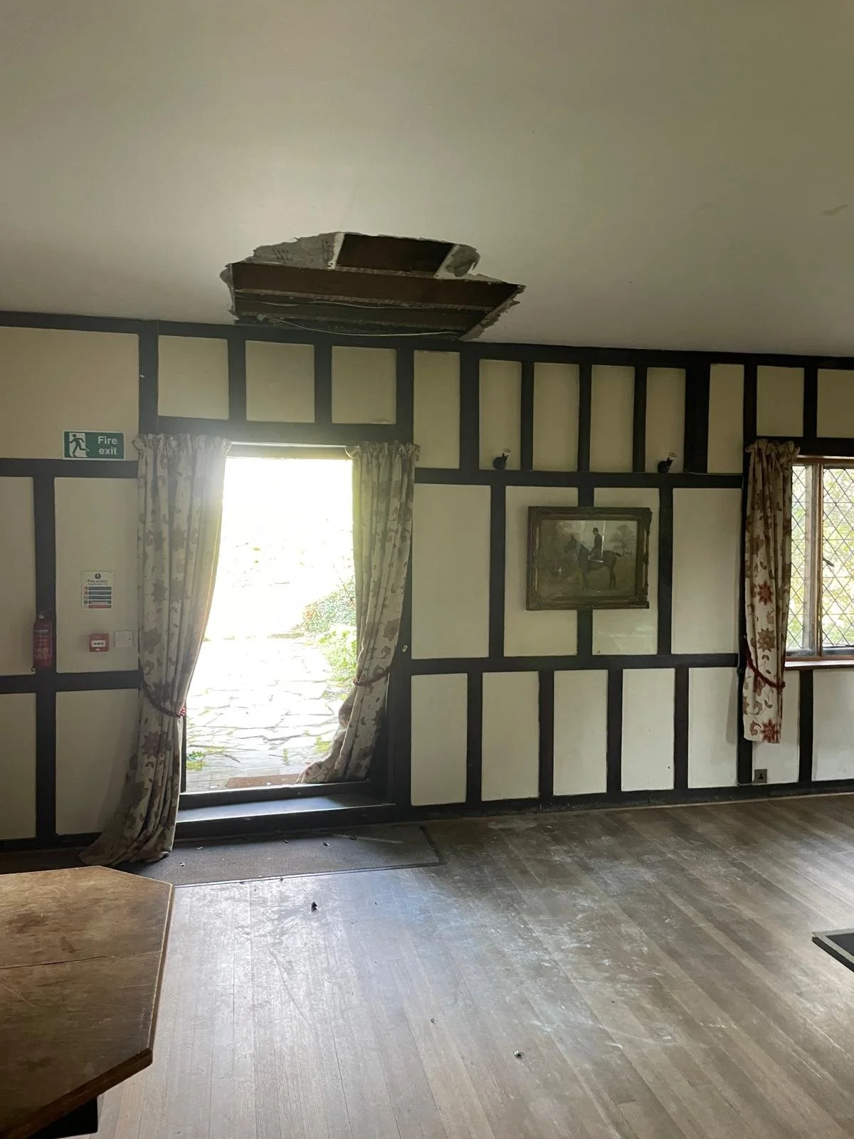 Room with damaged ceiling, open door to outside, patterned curtains, and framed artwork on wall.