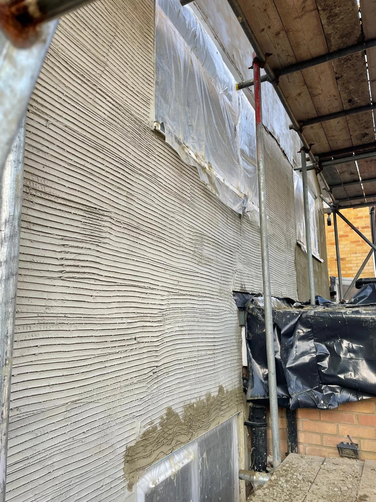 Exterior wall under construction with textured stucco, plastic sheeting, and scaffolding.