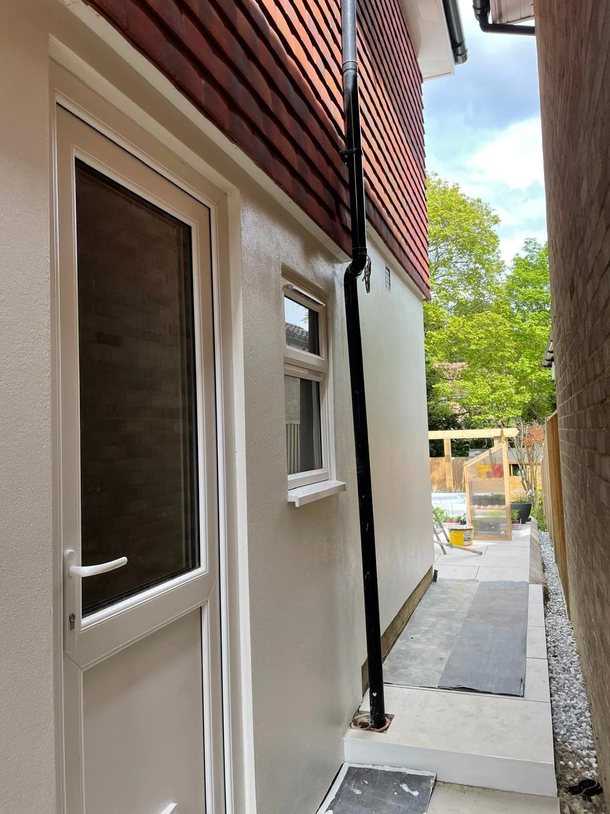 Side yard of a house showing a door, a small window, a downspout, and construction materials in the distance.
