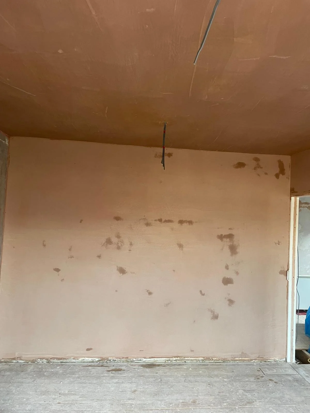 An unfinished room with a drywall wall showing patches of mud and an unpainted wooden ceiling. Electrical wires hang from the ceiling.
