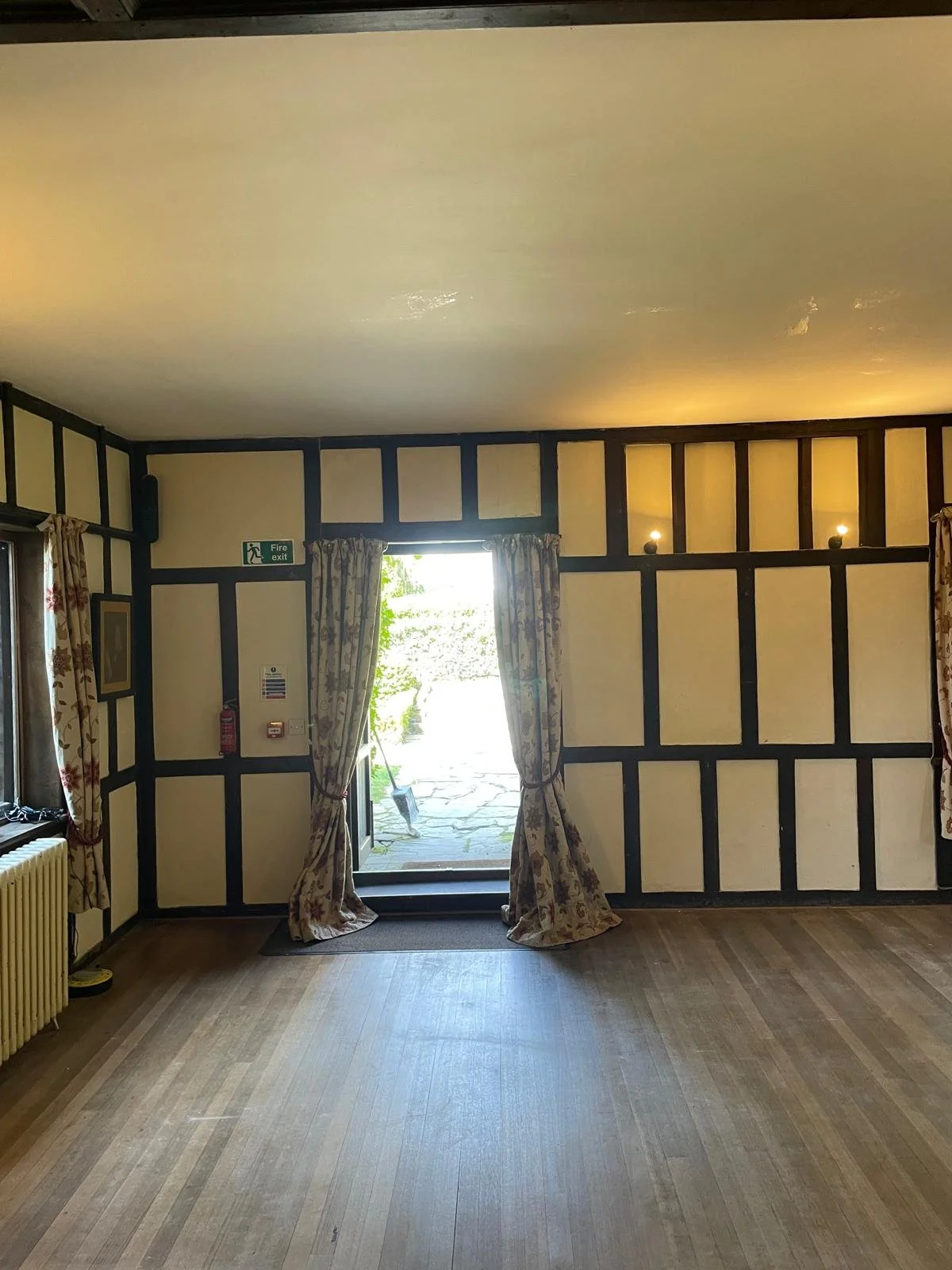 Empty room with wooden floor and cream-colored walls with dark trim, open door with floral curtains leading outside, window with similar curtains, fire exit sign, and wall-mounted lights.