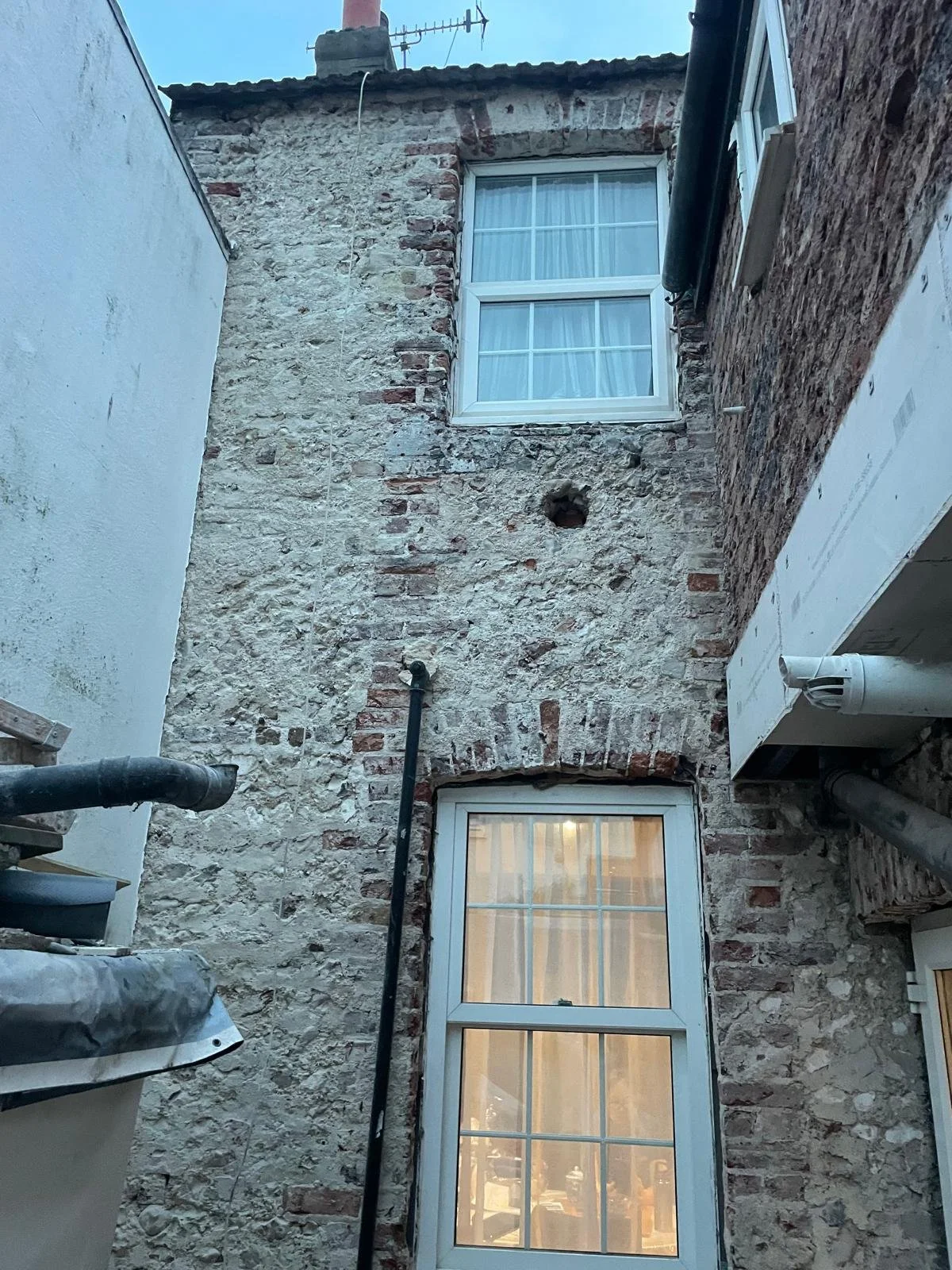 Back view of a narrow brick alley with two windows on a stone wall, pipes and an air vent visible.