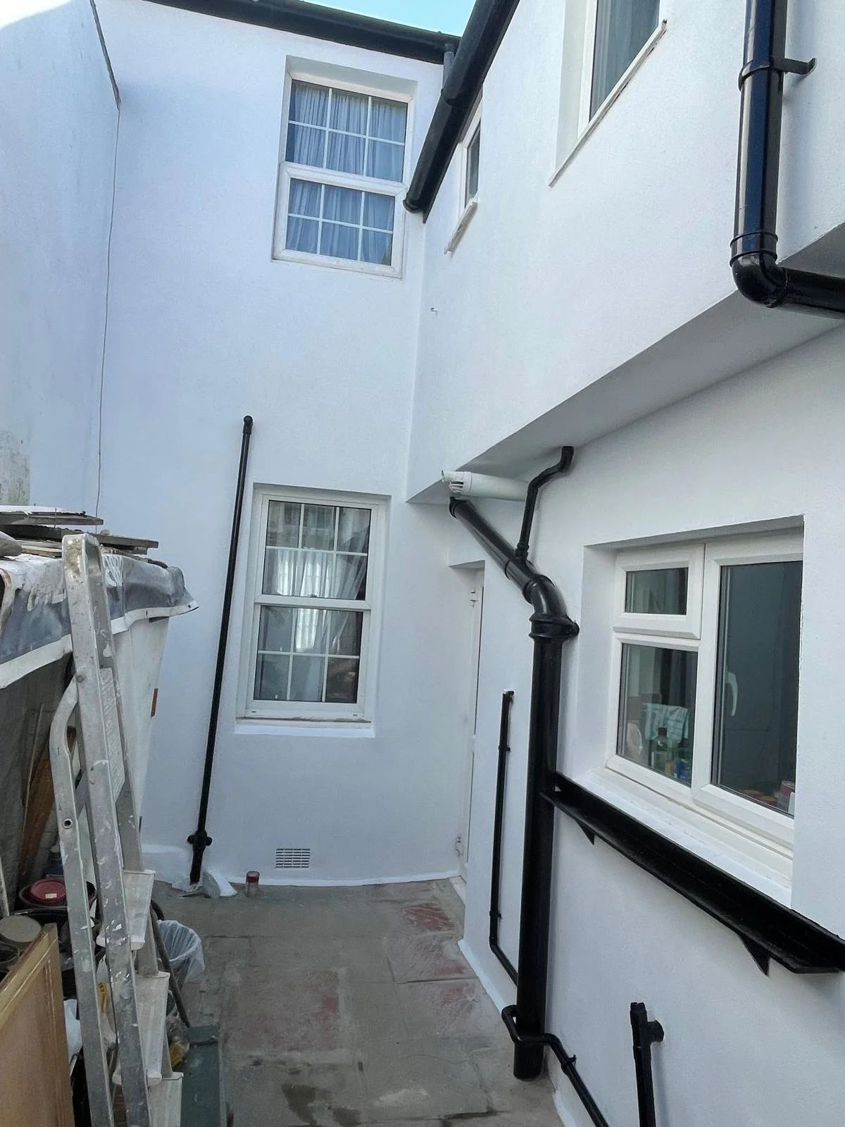 A small, enclosed outdoor area with white walls, two windows with curtains, and black drainage pipes attached to the wall.