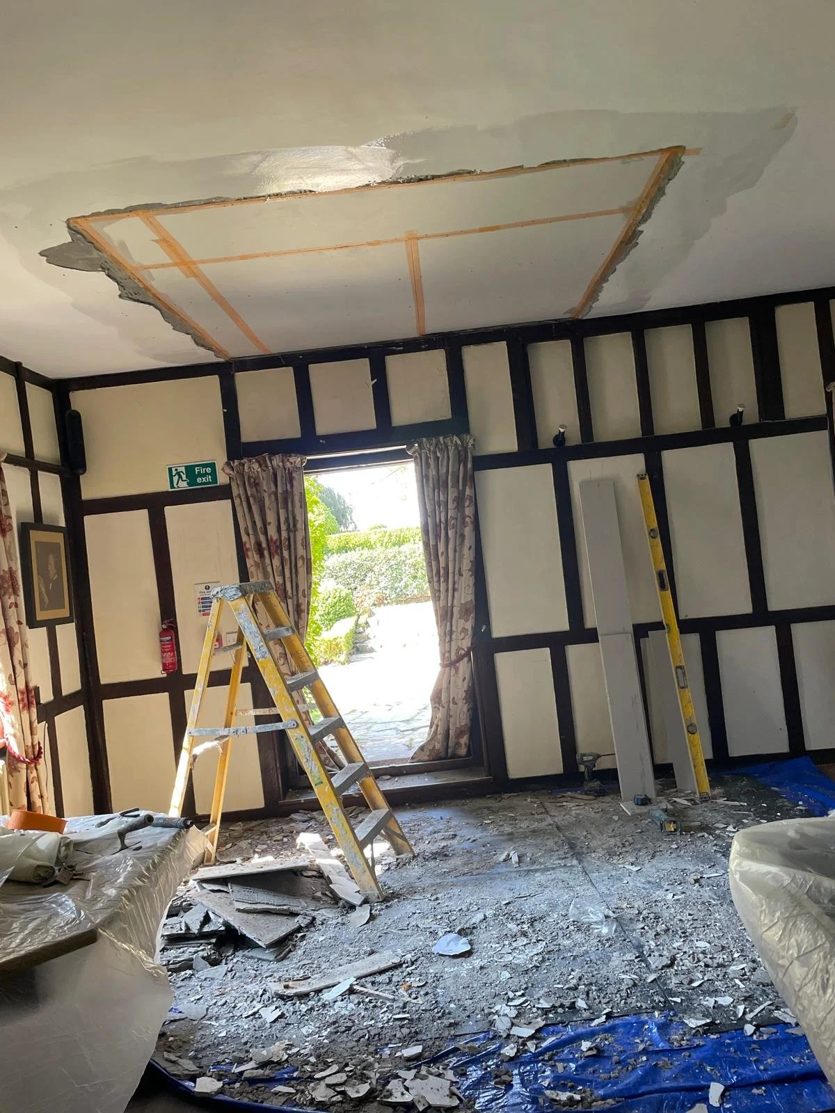 A room under renovation with a ceiling partially removed, revealing the structure above. Ladders and construction materials are present, and the floor is covered in debris.