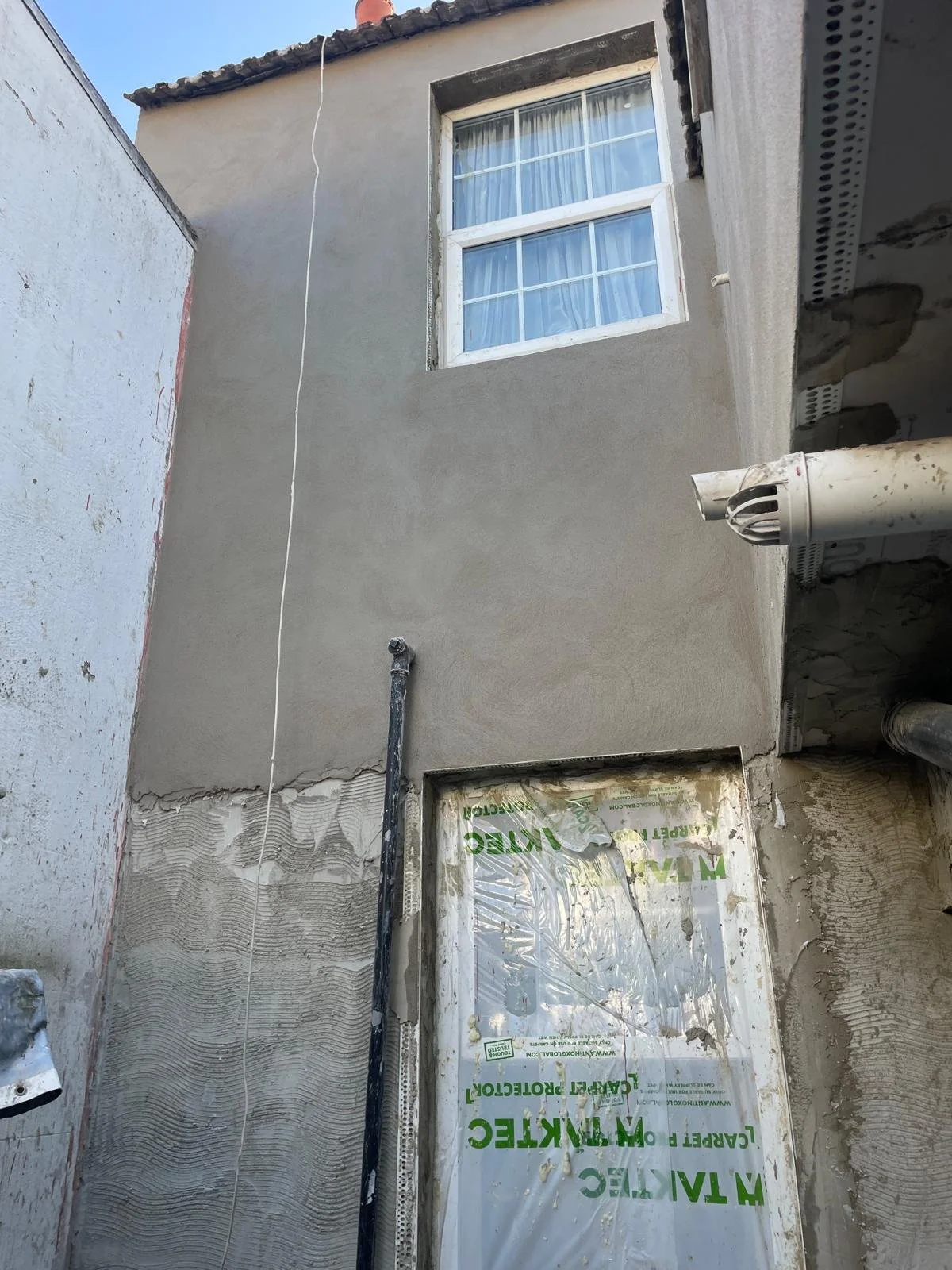 Building exterior under construction with a window, a drainpipe, and a wall with mixed unfinished and finished surfaces.