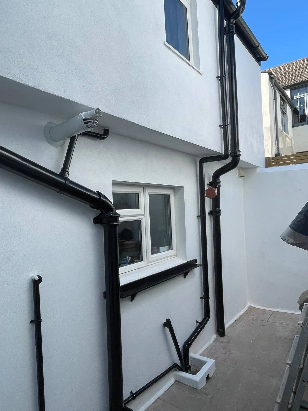 Exterior white building wall with new black drainage pipes running vertically and horizontally near a small window.