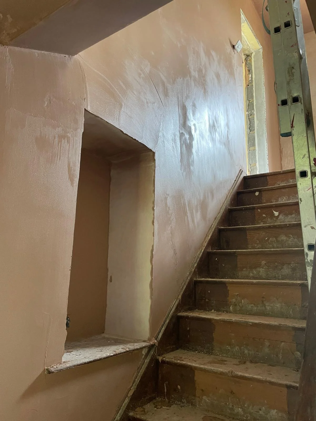 Interior of a house staircase undergoing renovation, with partially sanded wooden stairs, a level tool, and freshly painted peach-colored walls.