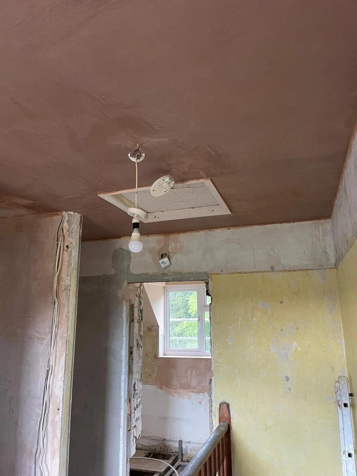 Room under renovation with unfinished walls, ceiling, and exposed wires. Temporary light bulb hanging from the ceiling, window letting in natural light.